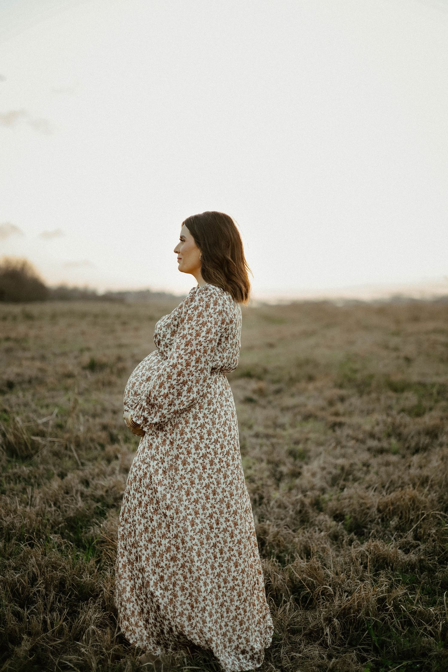 A silhouetted figure in a floral maxi dress stands in a grassy field at sunset, showcasing an expectant form.