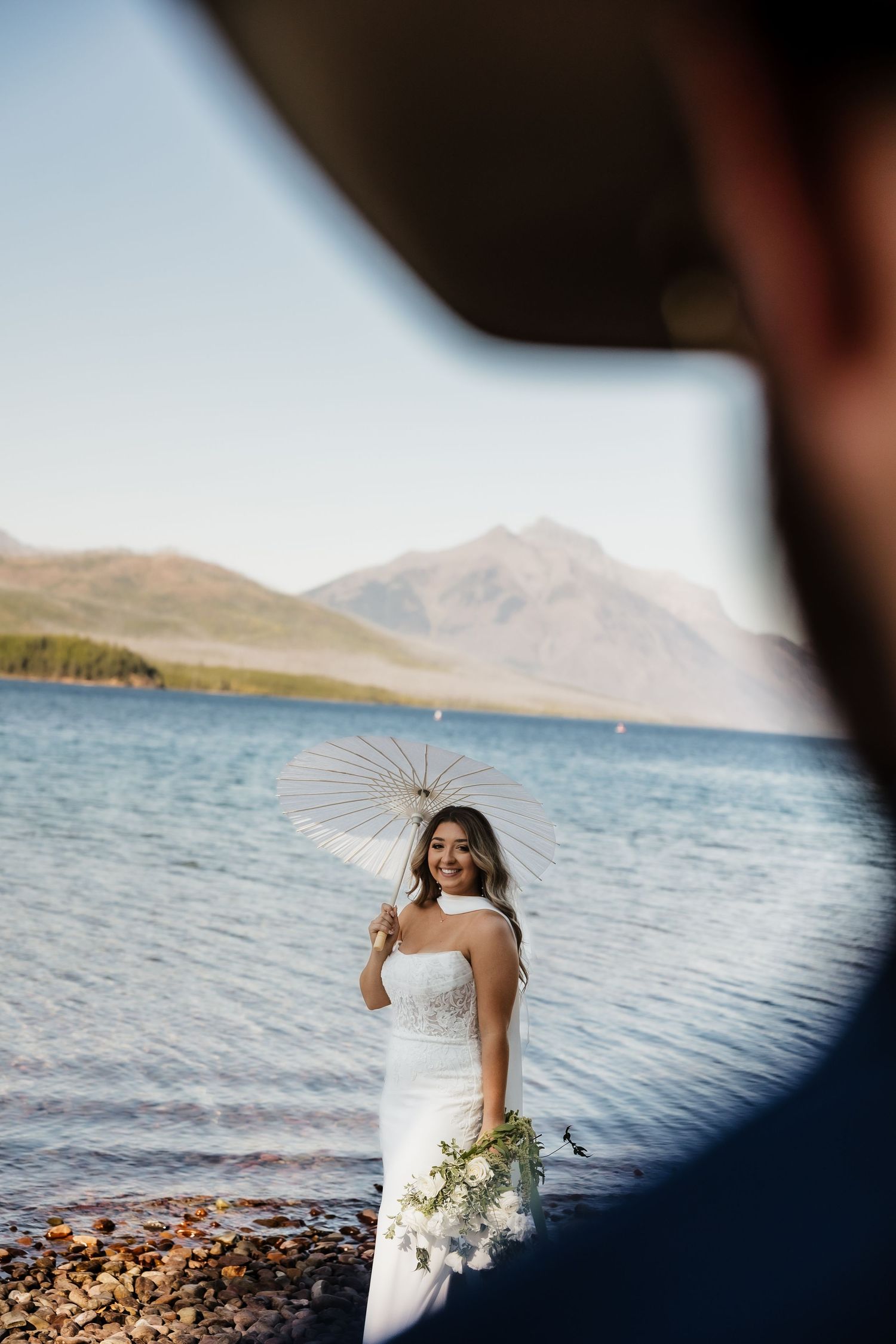 A coastal wedding scene with a white dress and parasol against a backdrop of mountains and sparkling water.