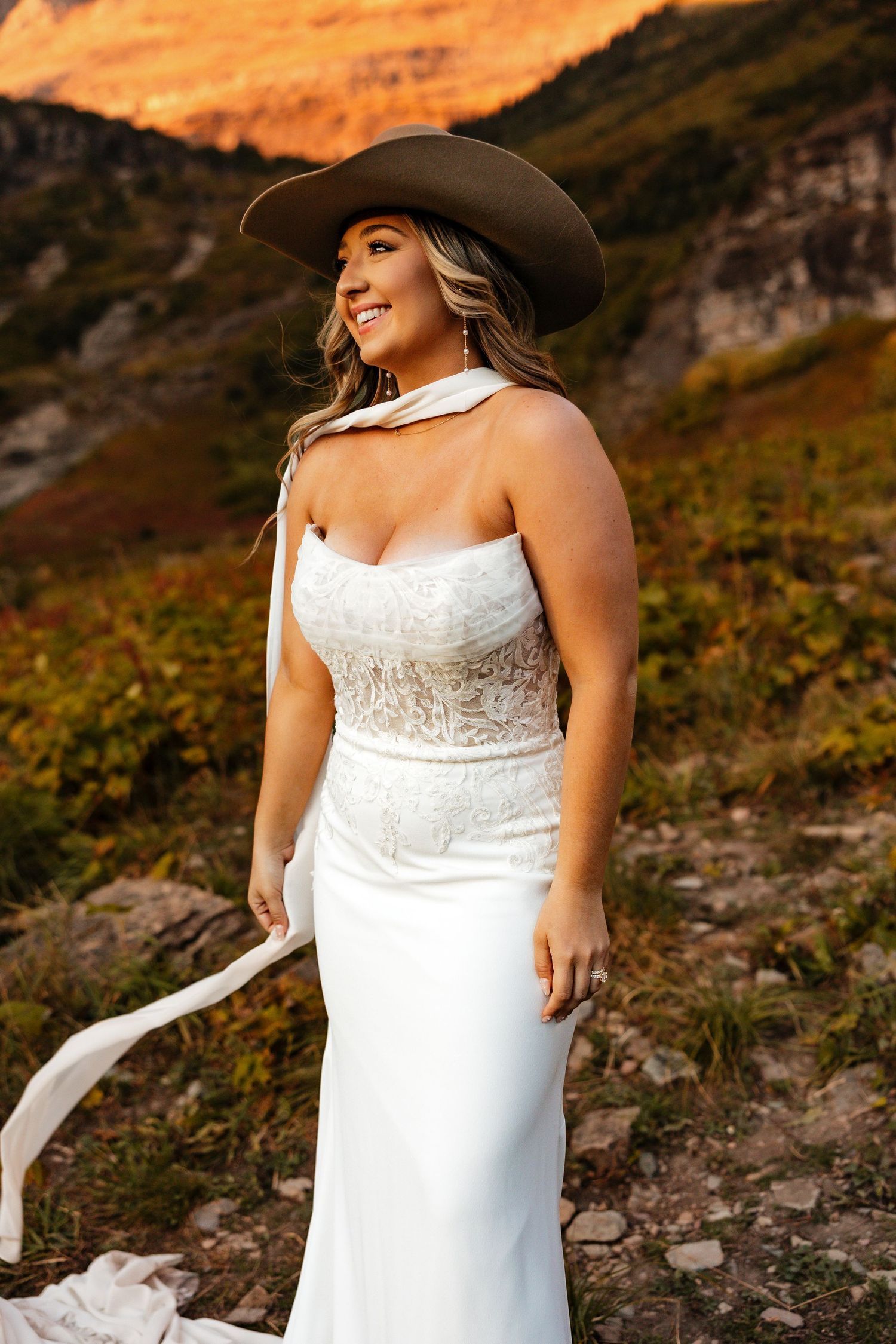 A bride in a fitted white gown and cowboy hat poses against scenic mountain backdrop at sunset.