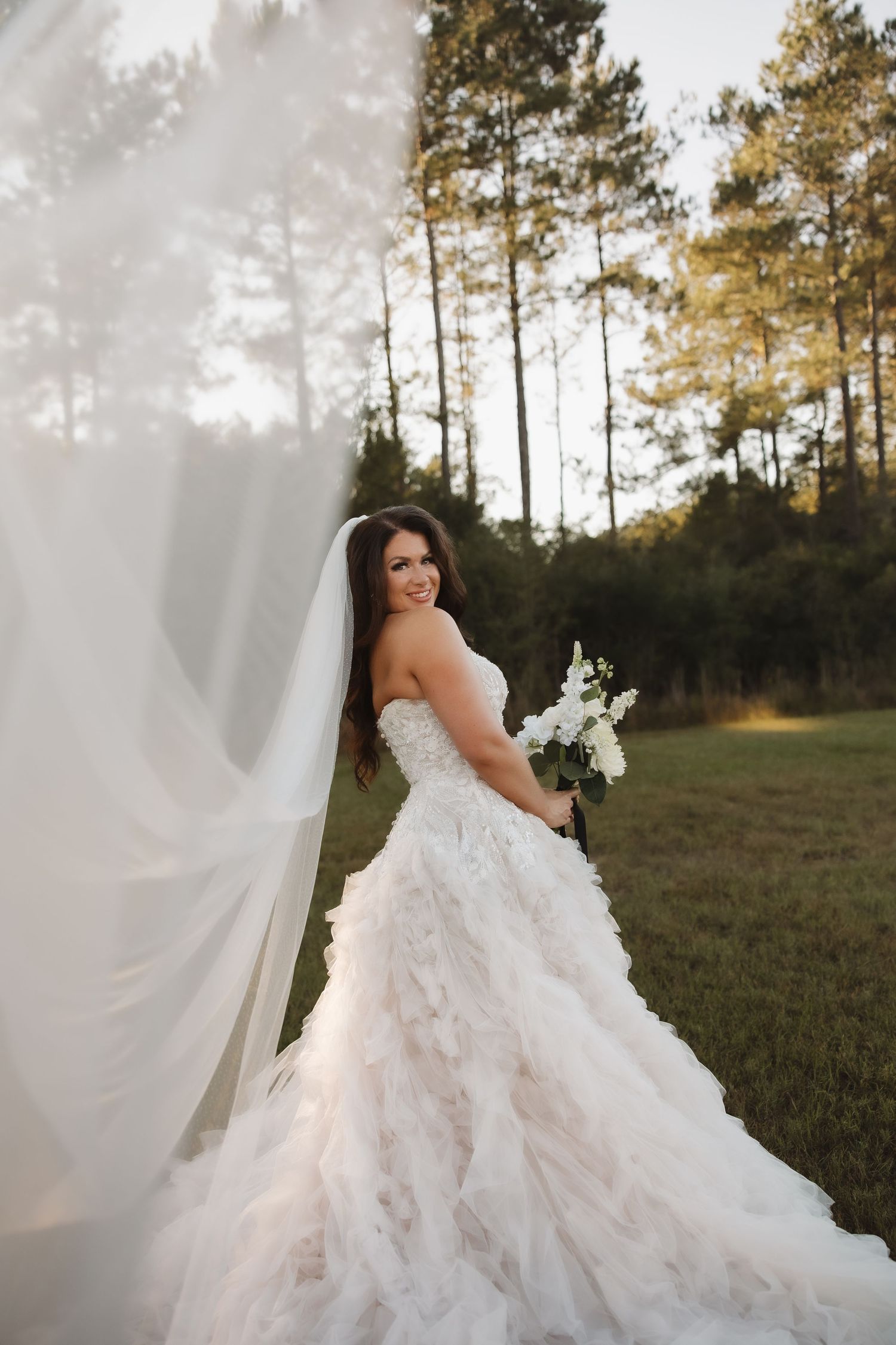 A bride in a flowing white ruffled wedding gown poses gracefully against a backdrop of tall pine trees at sunset.