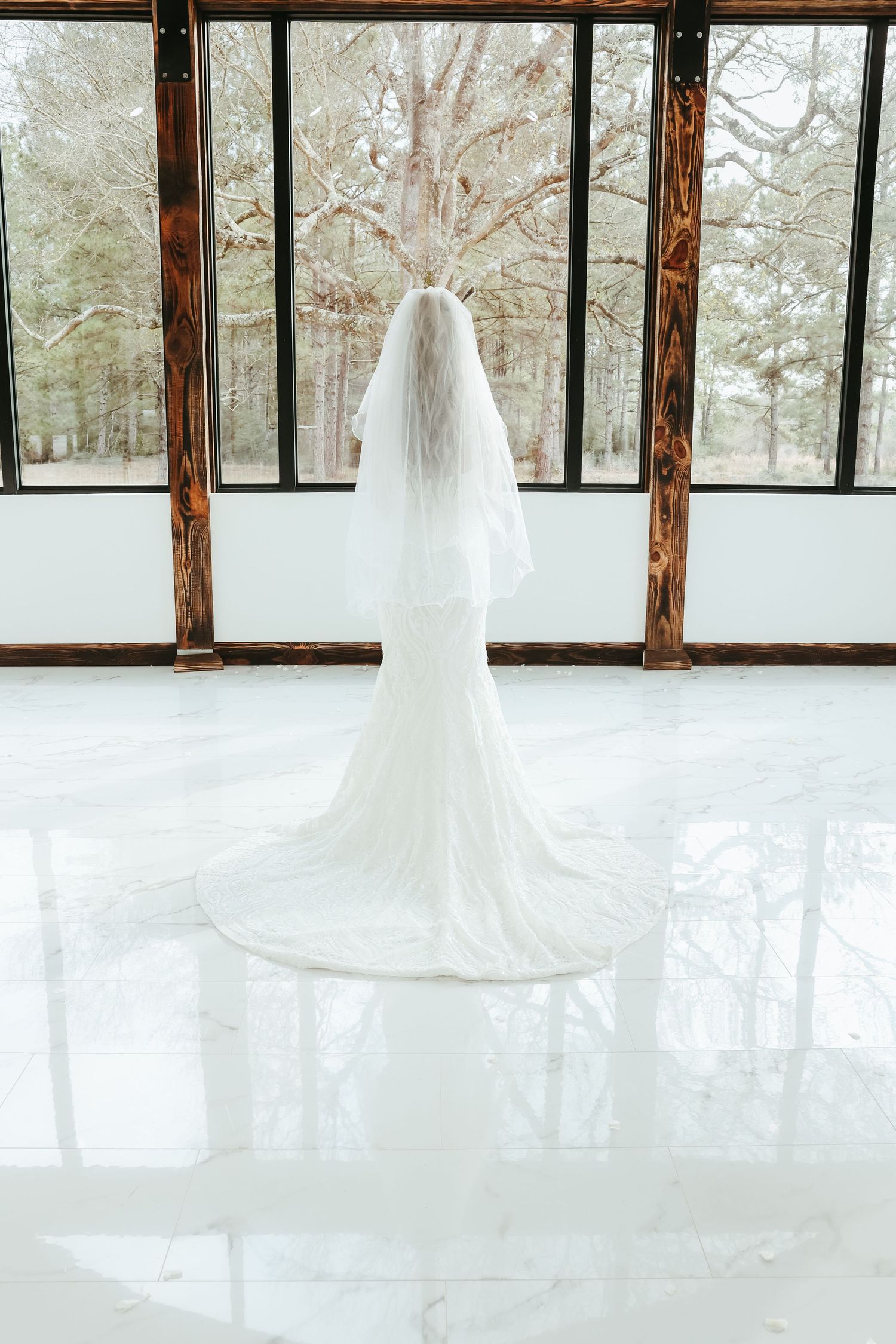 A flowing white wedding dress with long veil stands elegantly against floor-to-ceiling windows overlooking a snowy forest.