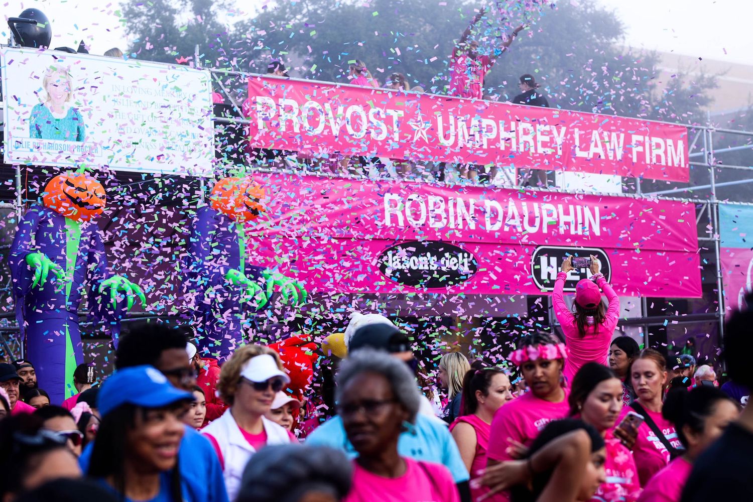 Crowd celebrates at outdoor event with pink banner and confetti during breast cancer awareness fundraiser or charity run.