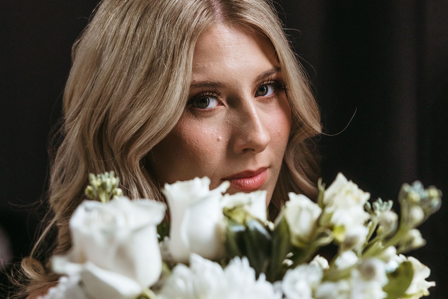 A person with blonde hair poses with a bouquet of white roses in soft dramatic lighting.