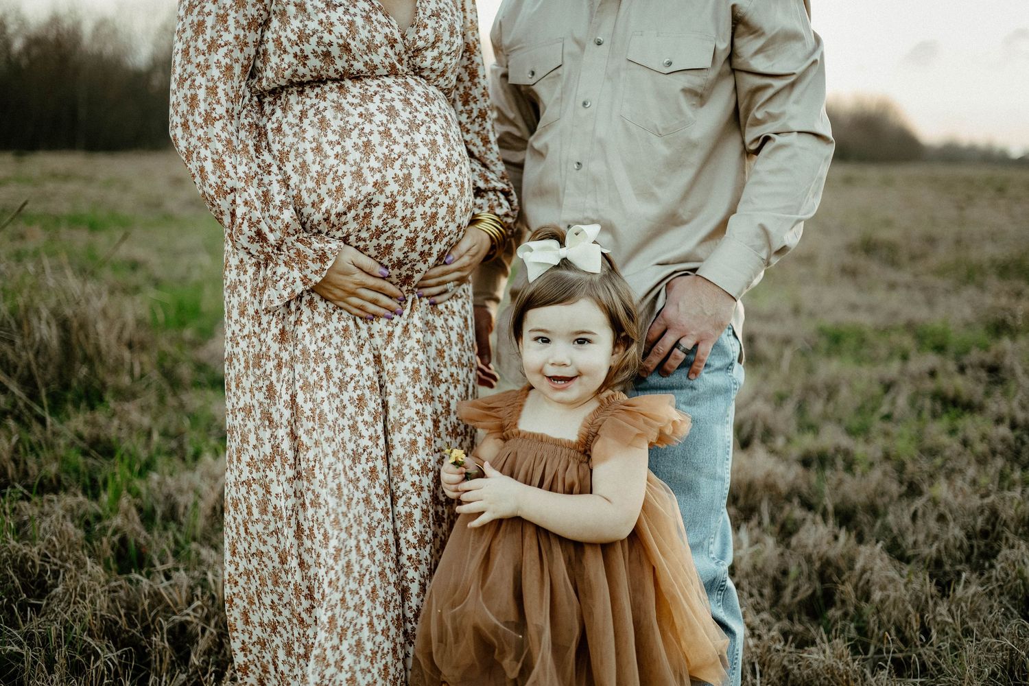 Family standing together in a rustic field wearing brown and beige tones during maternity photo session.