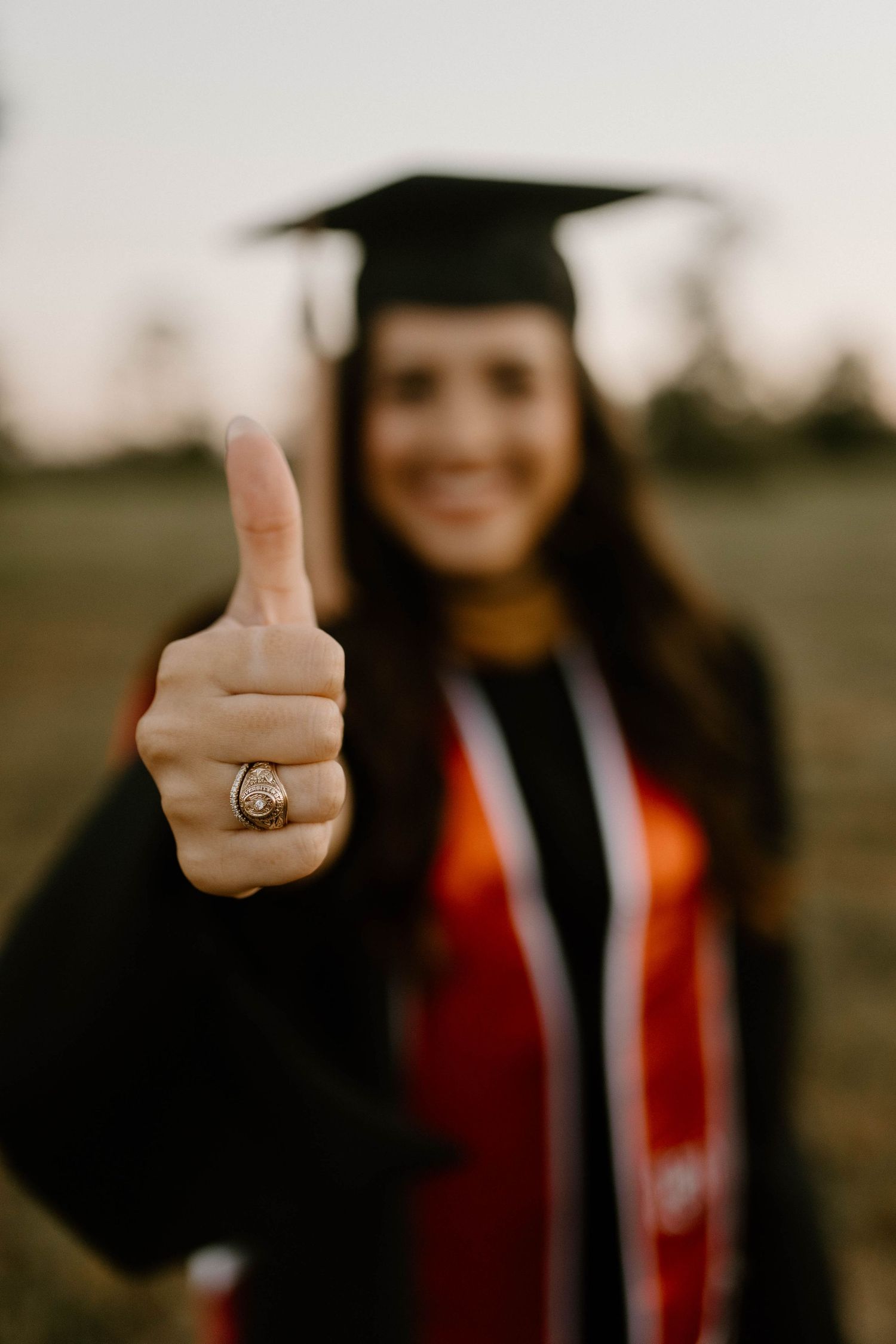 A graduate in cap and gown gives a thumbs up while wearing a class ring outdoors at sunset.
