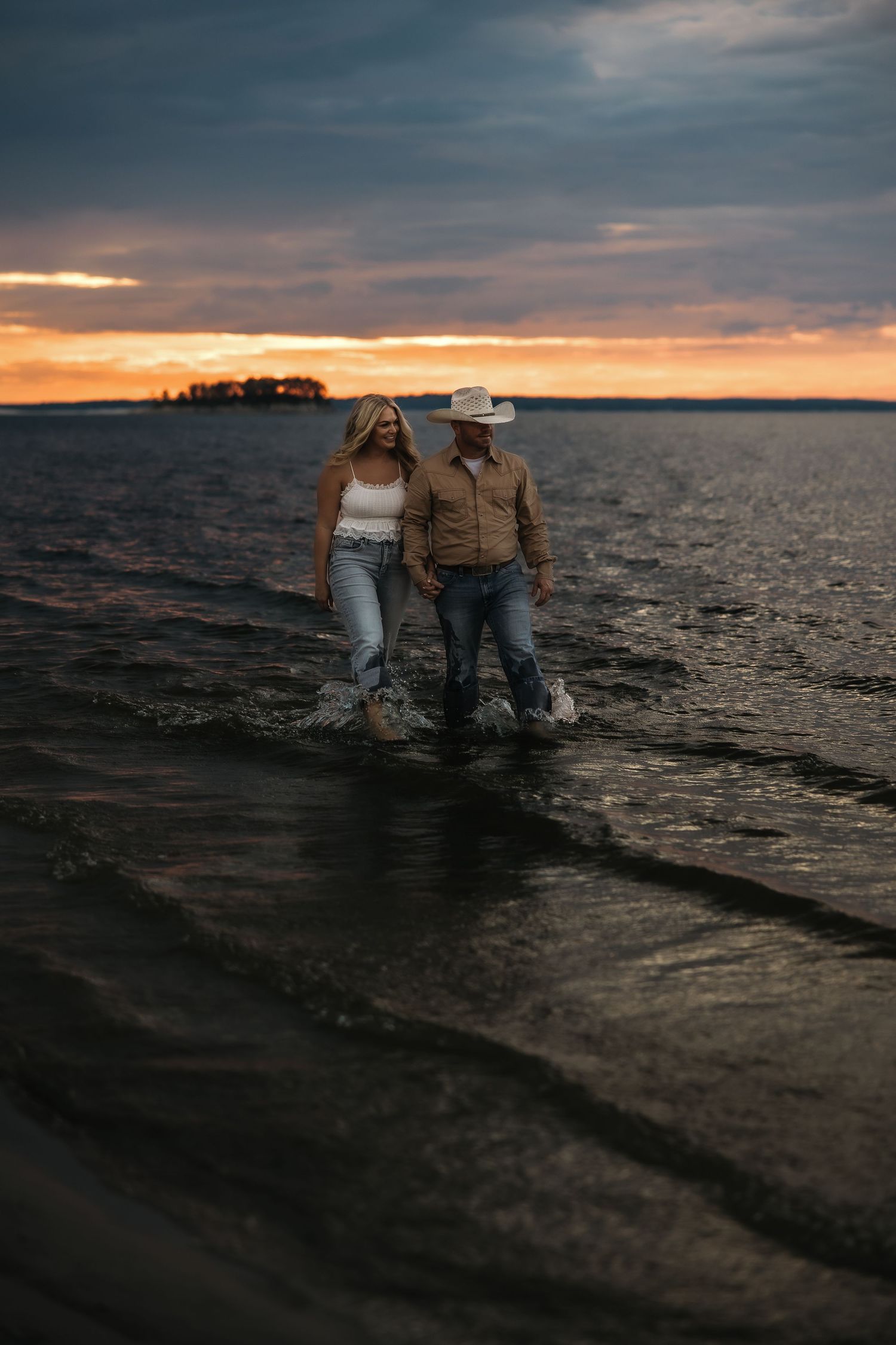 Two people sitting together on a rocky shoreline at sunset with dramatic cloudy skies over the water.
