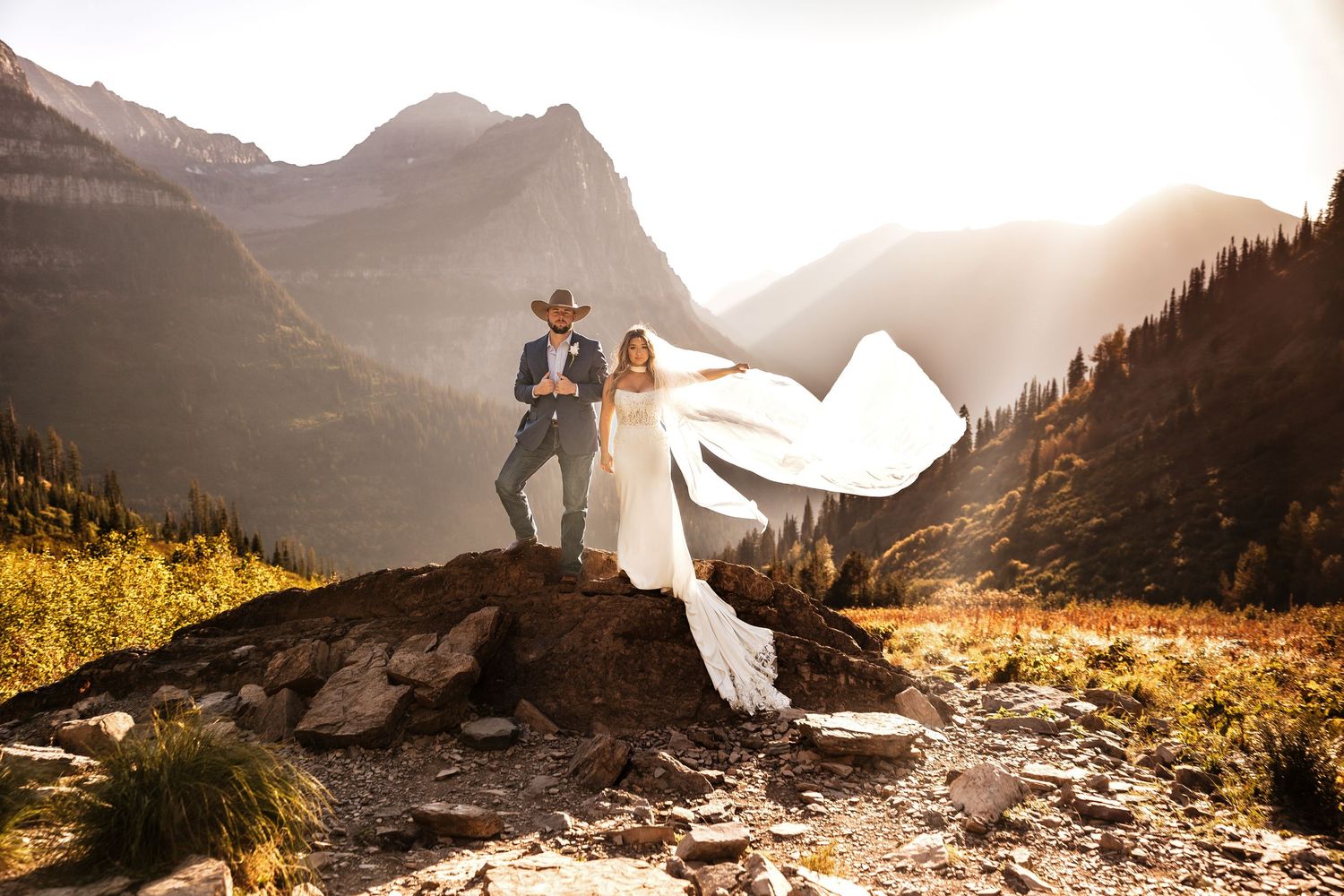 Couple stands on mountain peak at sunset with flowing white dress catching wind in dramatic mountain landscape.