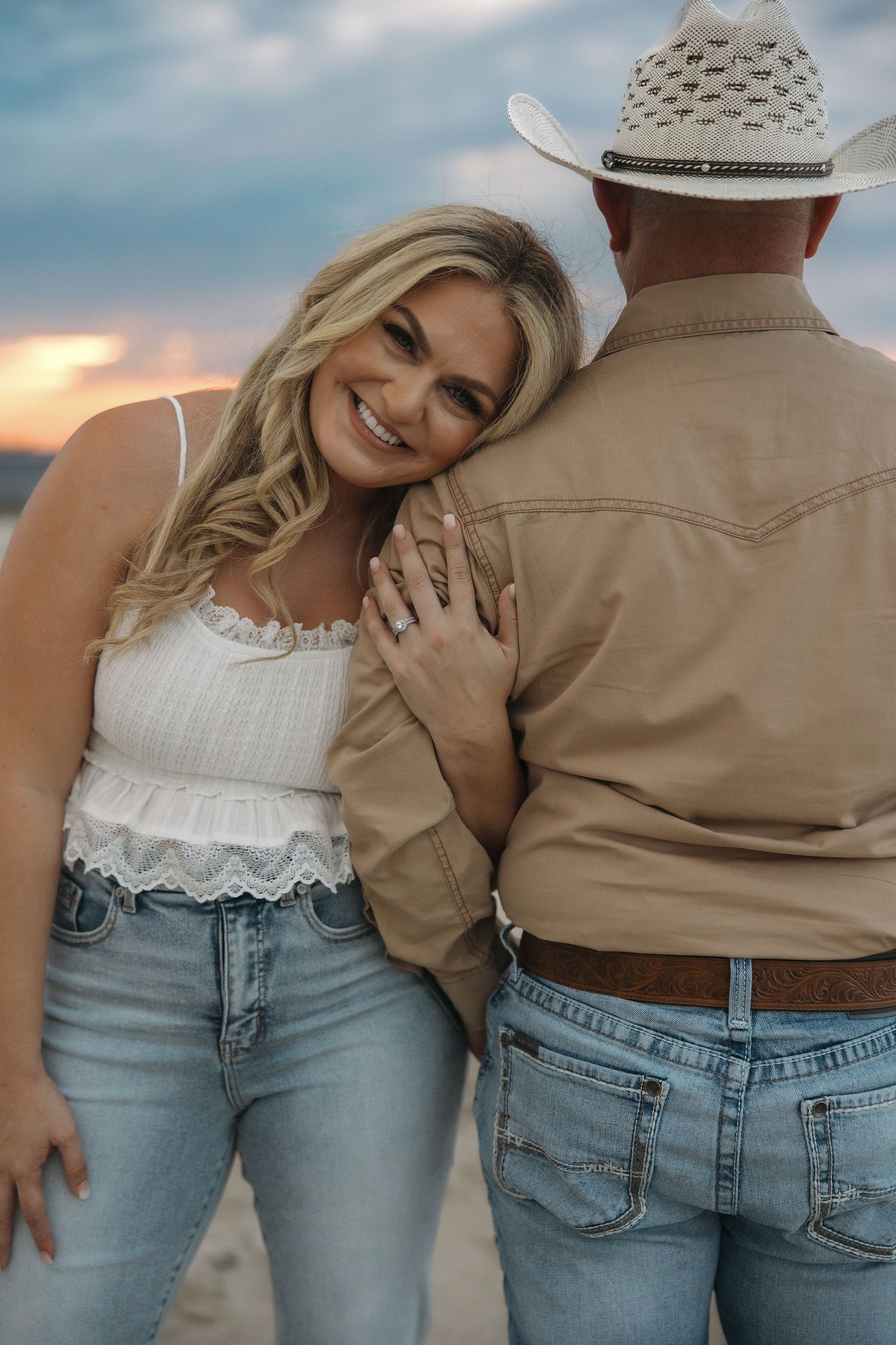 A couple in casual attire embracing against a sunset sky, with one wearing a white top and jeans.