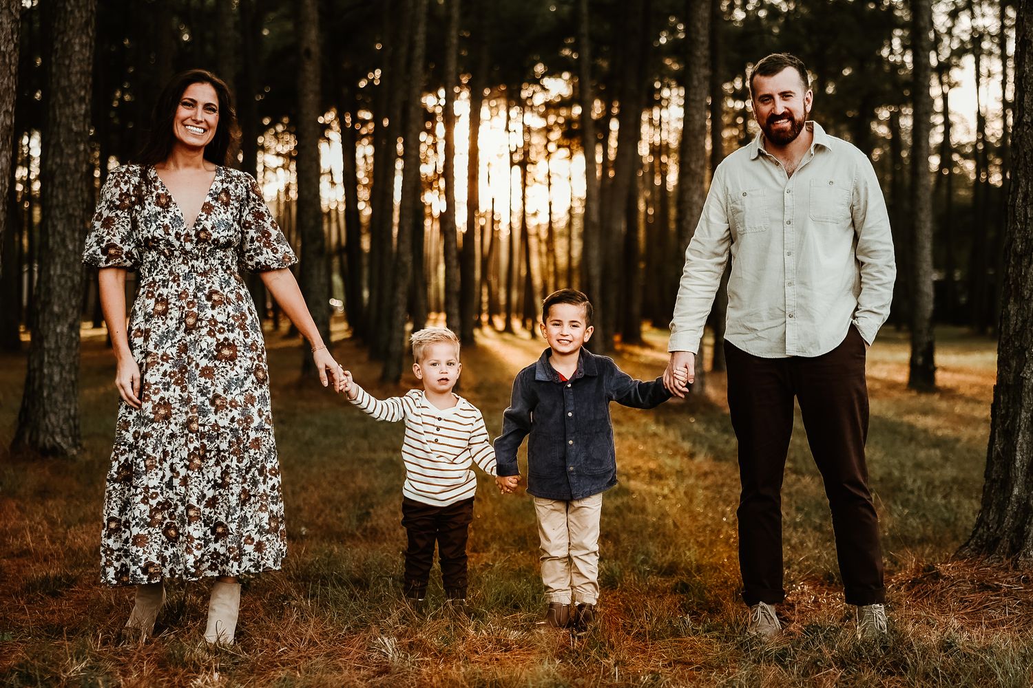 A family stands together in a sunlit pine forest at golden hour wearing coordinated casual outfits.