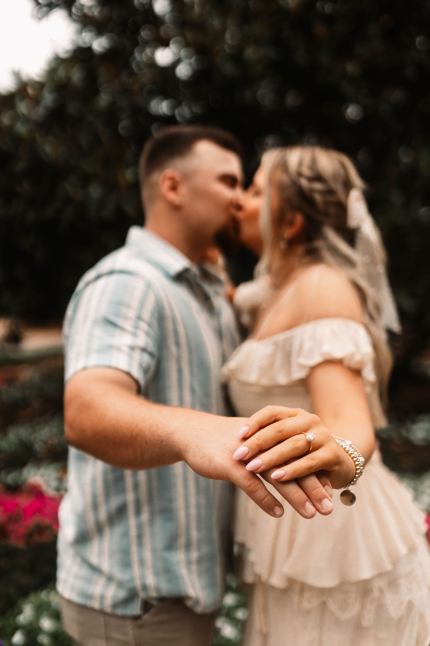 Close-up of hands showing engagement ring while couple shares romantic moment in garden with flowers.