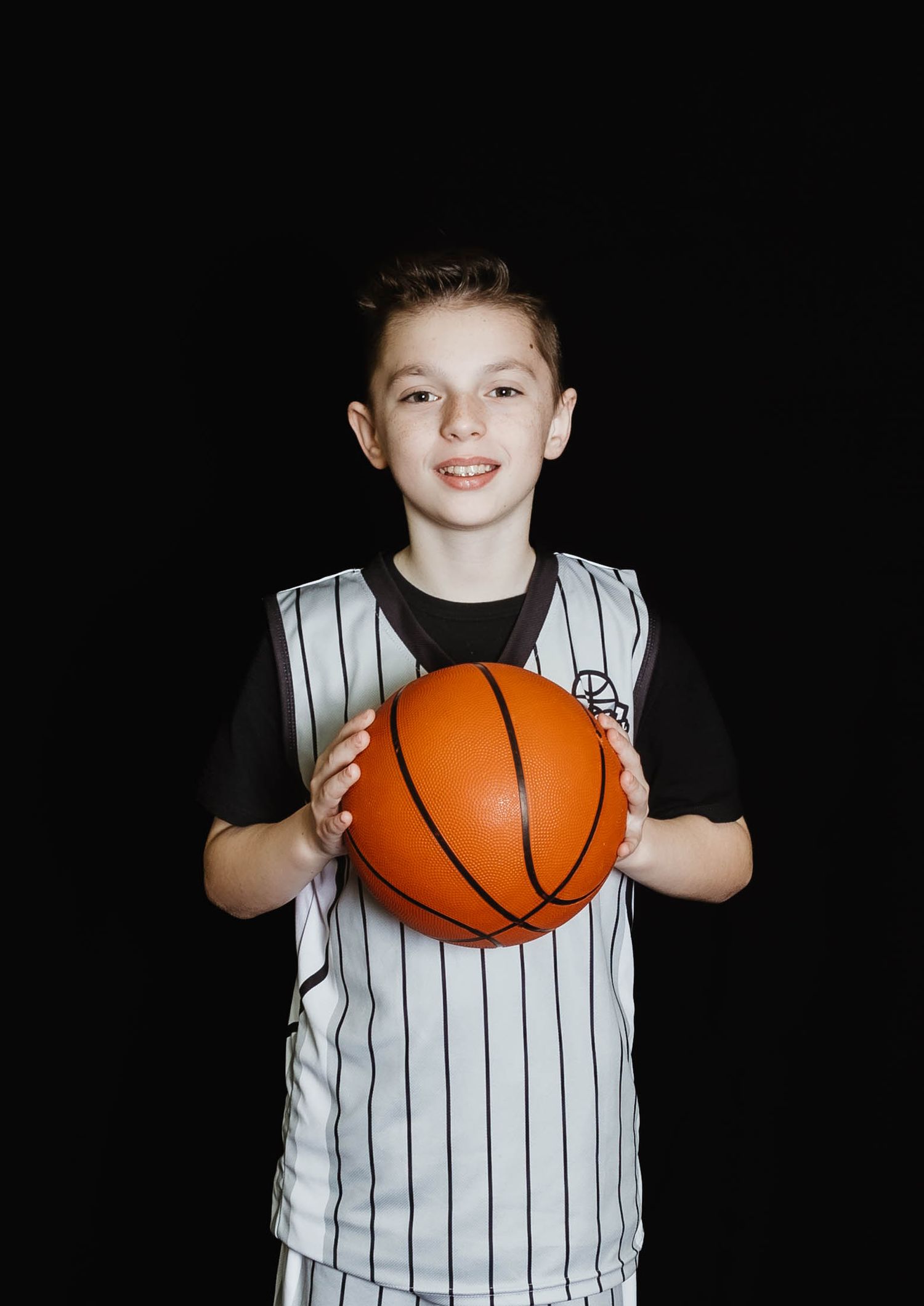 Young basketball player wearing white pinstriped jersey holds orange basketball against dark background.