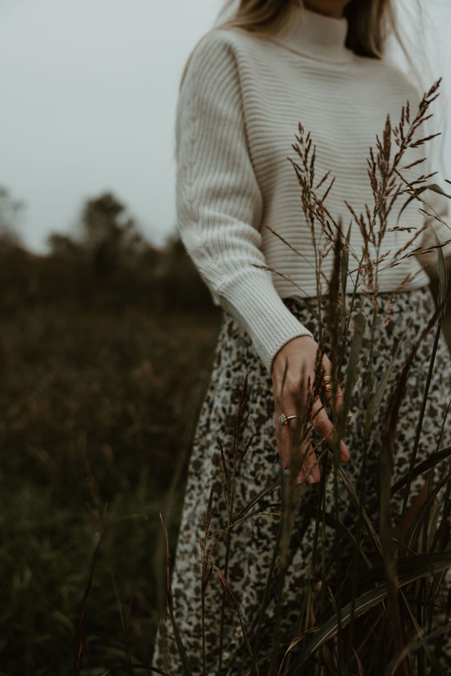 Moody autumn fashion featuring cream sweater and floral skirt among tall dried grasses in dark field.