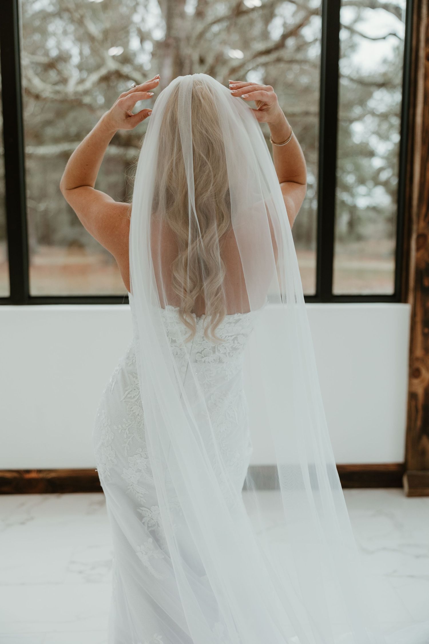 Back view of bride in white lace wedding gown adjusting her long flowing veil against a window backdrop.