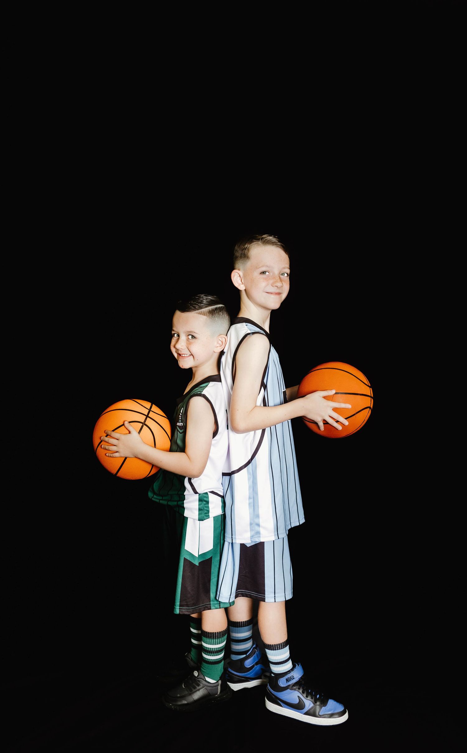 Two young basketball players in uniforms stand back-to-back holding orange basketballs against a black background.