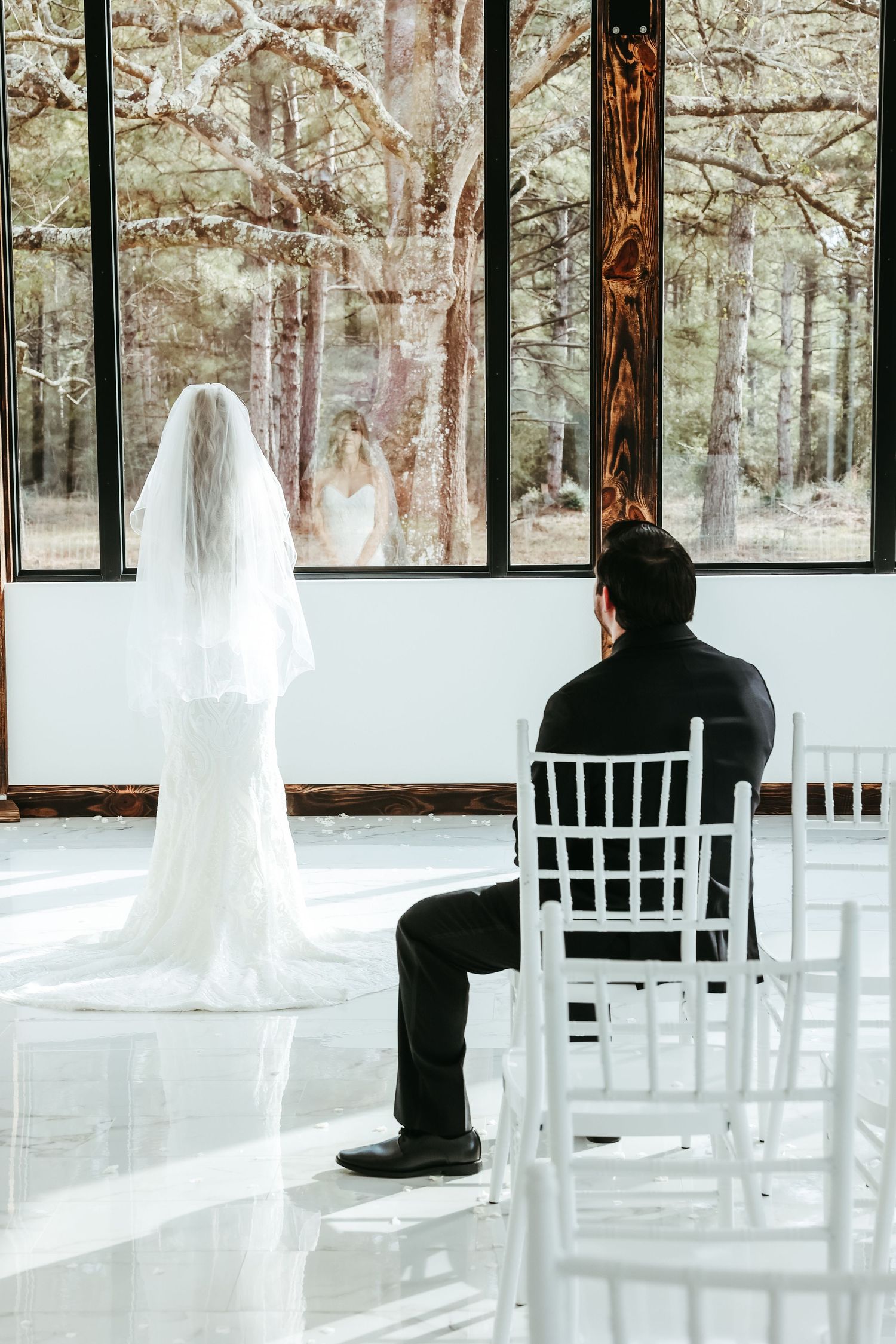 A bride in a flowing white veil stands before large windows in a modern wedding venue while someone sits in a white chair.