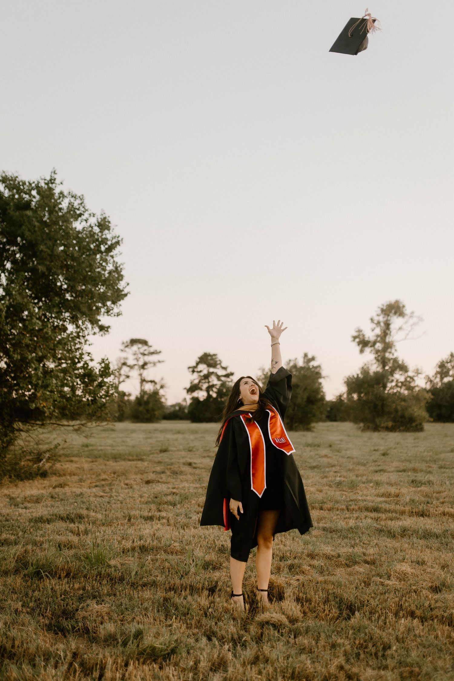 A student in graduation regalia throws their cap in a grassy field at sunset with trees in the background.