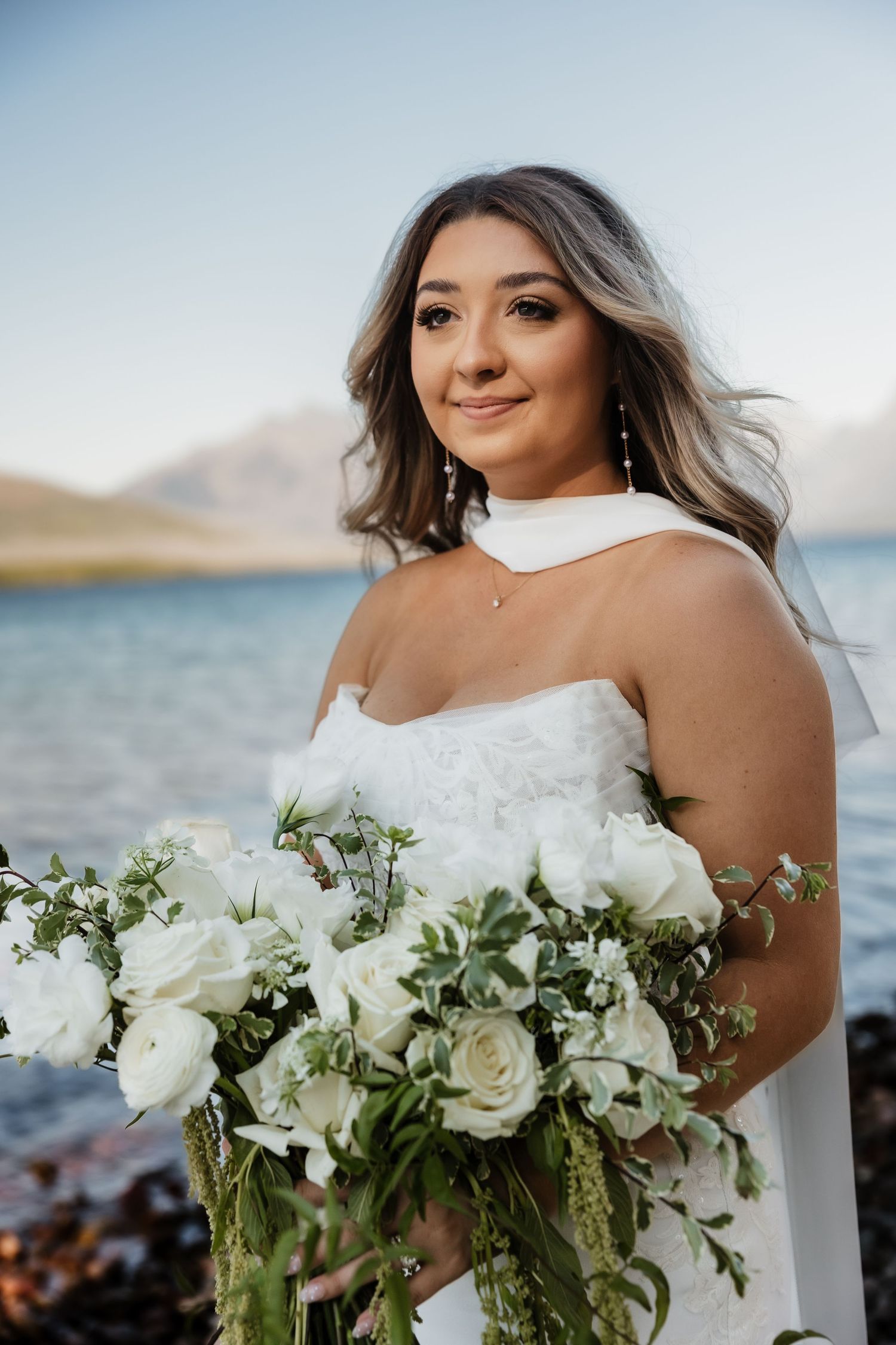 A bride in a white off-shoulder gown holds a lush bouquet of white roses and greenery by the ocean.