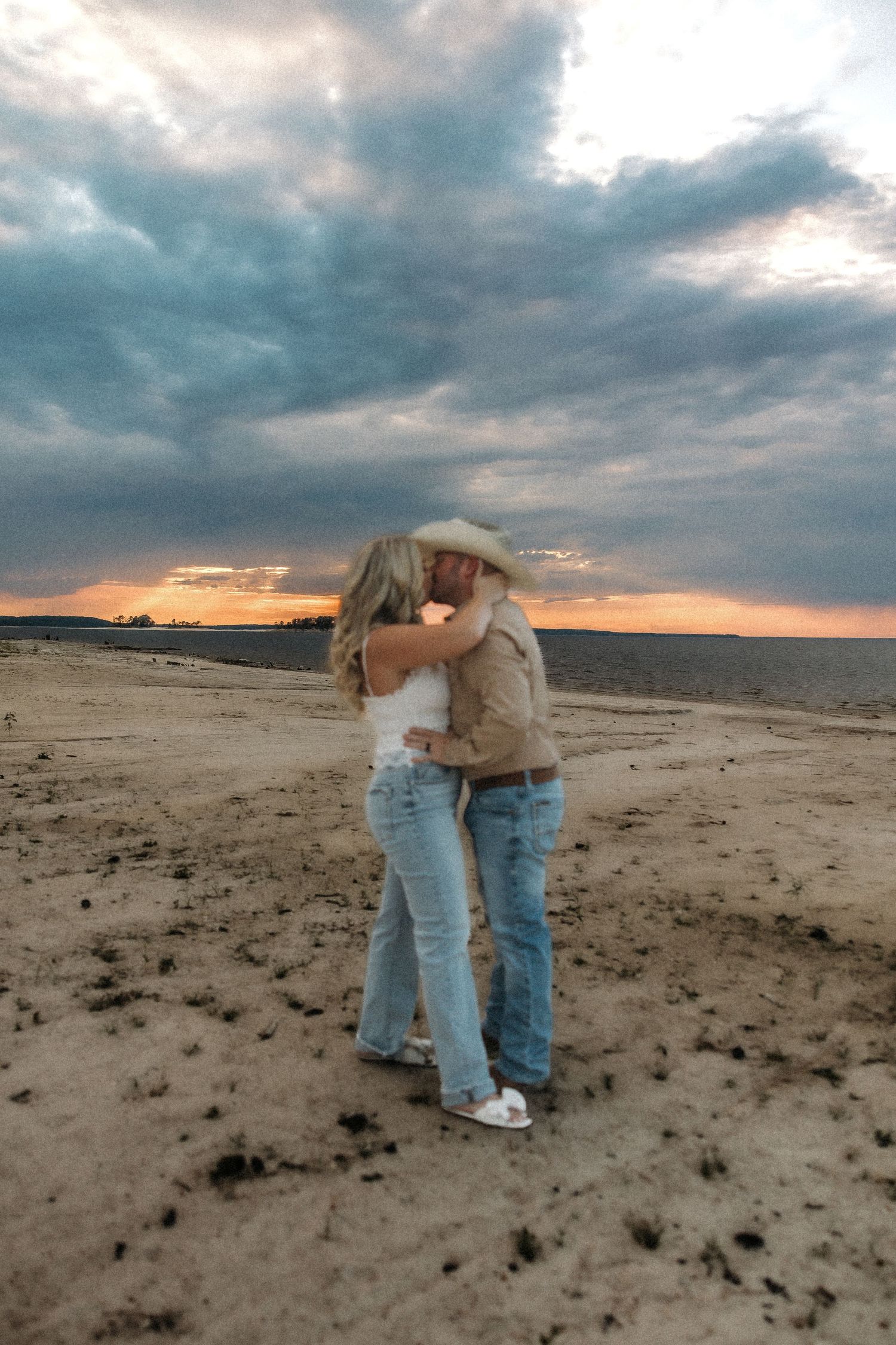 A couple embraces on a desert plain under dramatic cloudy skies at sunset.