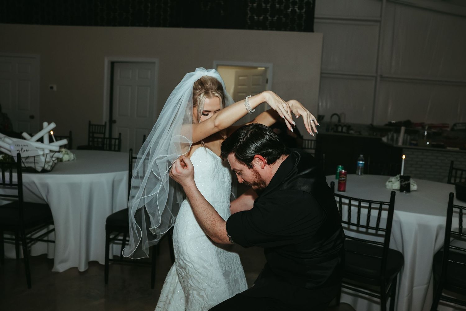 A bride in a white gown and veil playfully dances with someone in a dark suit at a wedding reception.