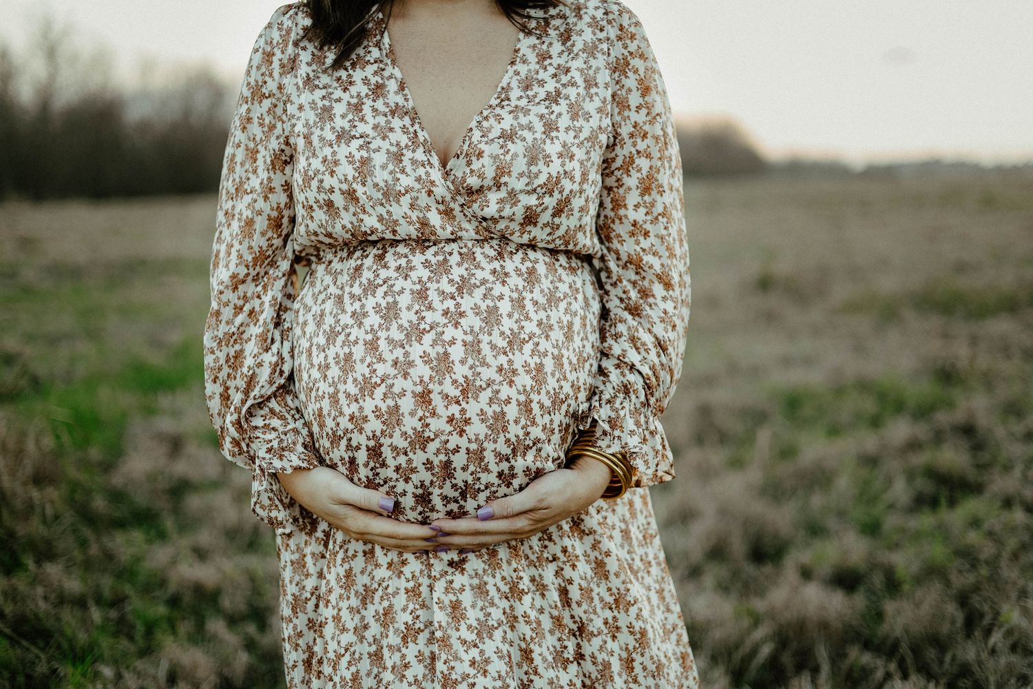 Pregnant woman in floral dress cradles baby bump while standing in rustic field at sunset.