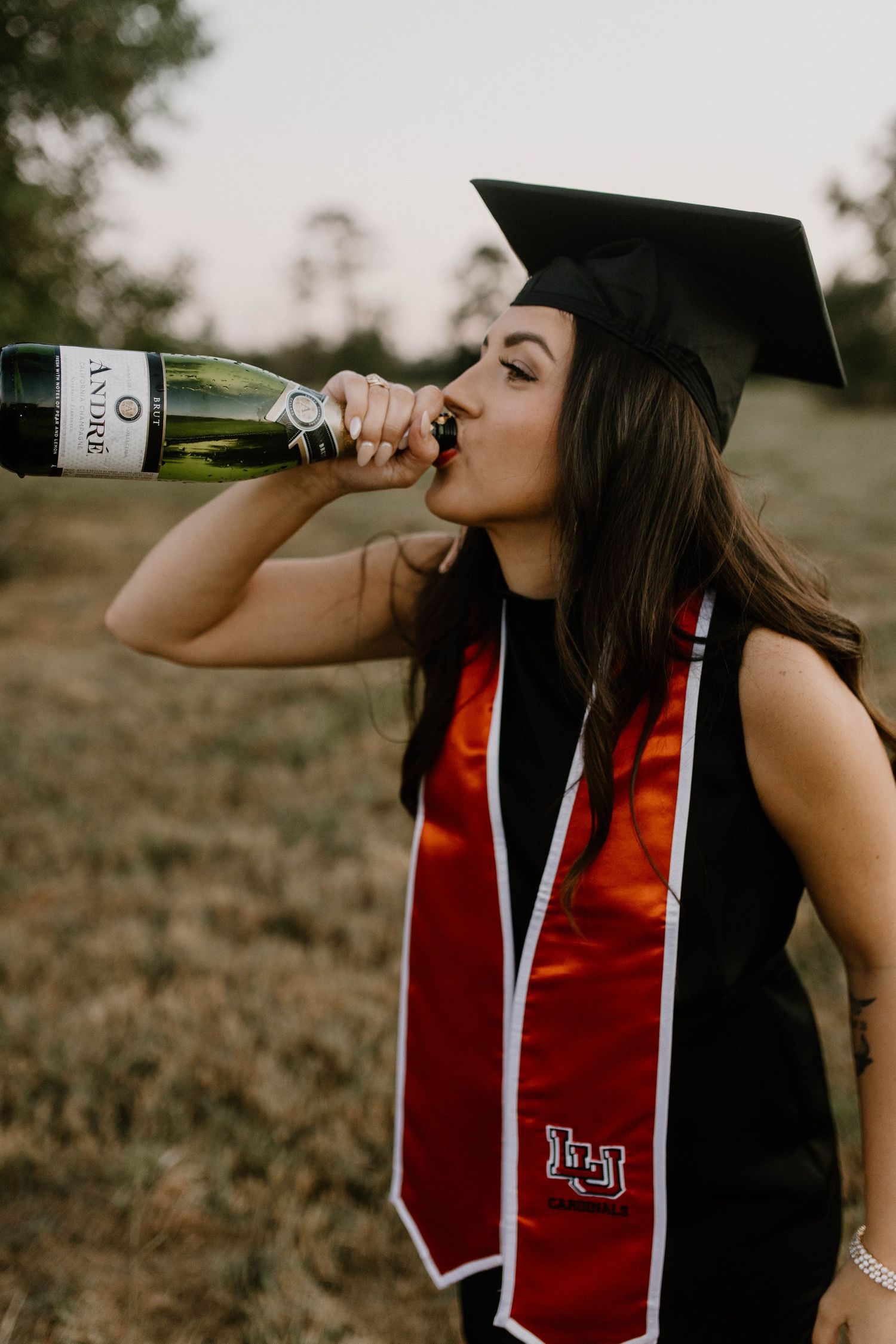 Graduate in academic regalia with red stole drinks from champagne bottle during graduation celebration photoshoot.