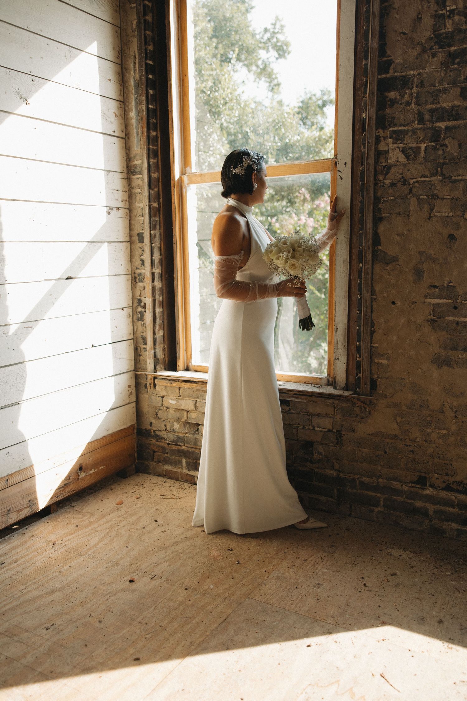 A bride in a white wedding dress stands by a rustic brick window, creating an elegant silhouette in natural light.