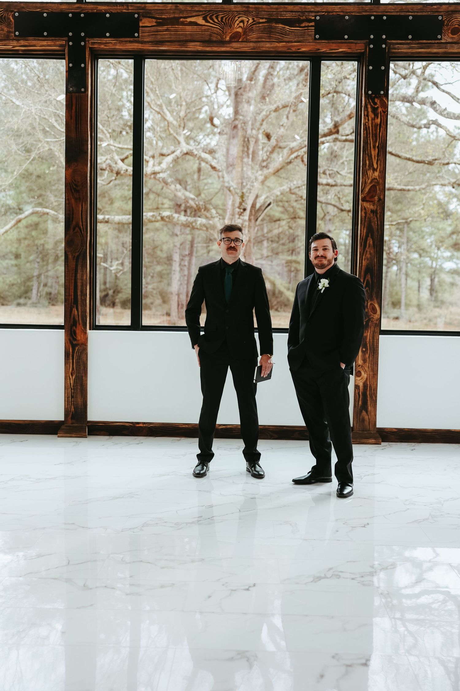 Two people in black formal attire stand in front of large windows in a bright, modern interior space with wood beams.
