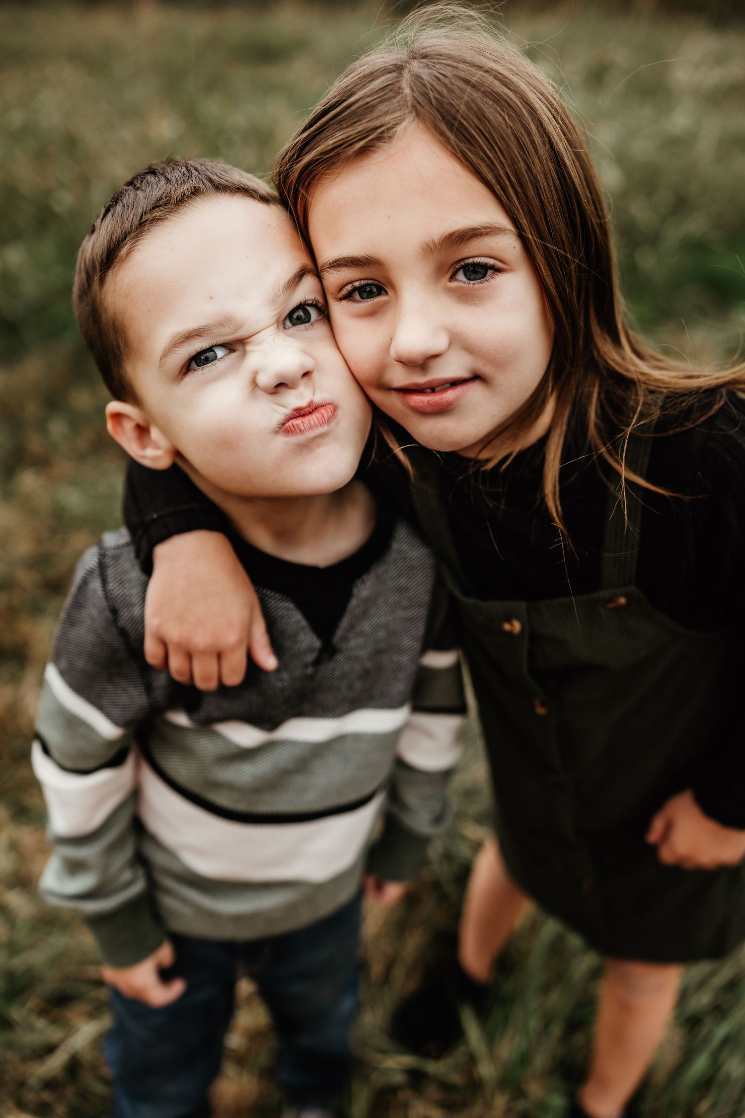 Two young children pose together outdoors, one making a silly face while wearing a striped sweater.