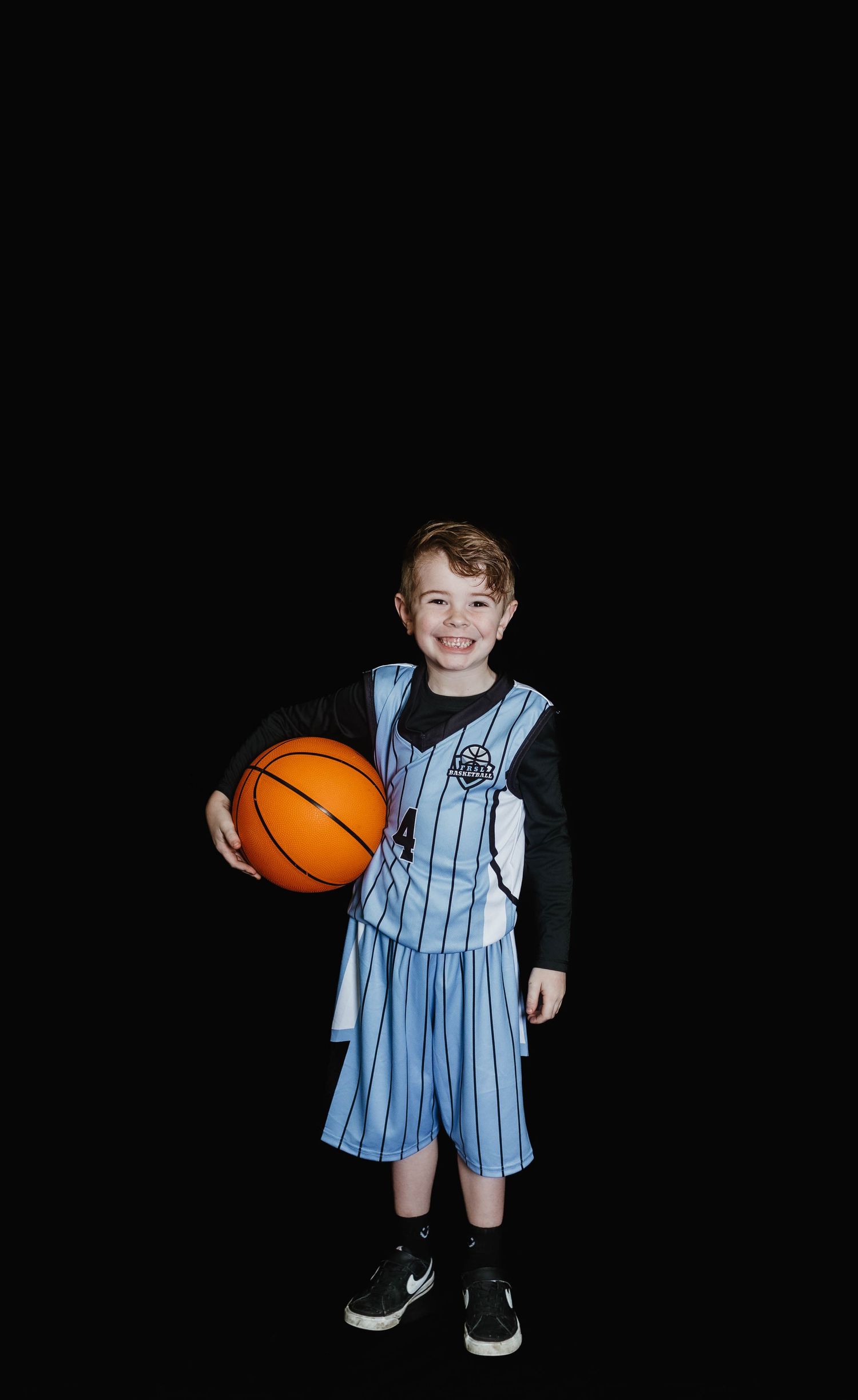 A young basketball player poses in light blue striped uniform while holding an orange basketball against black background.
