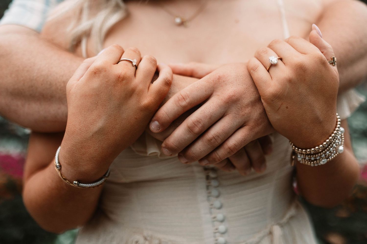 Close-up of hands with rings and bracelets embracing around a white corset dress.