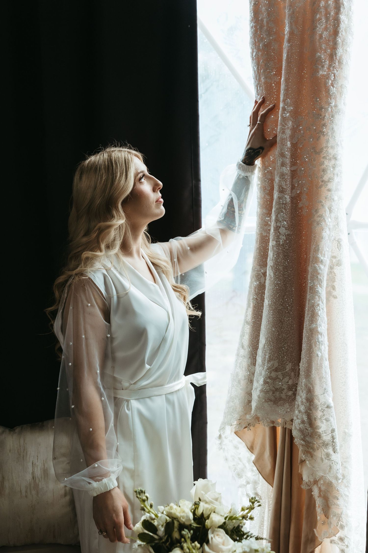 A person in a white robe stands near a window with sheer curtains while holding a bouquet of white flowers.