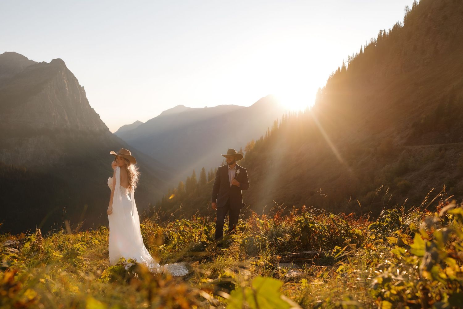 Couple poses in golden wildflower meadow with sunlit mountain peaks in background during sunset.
