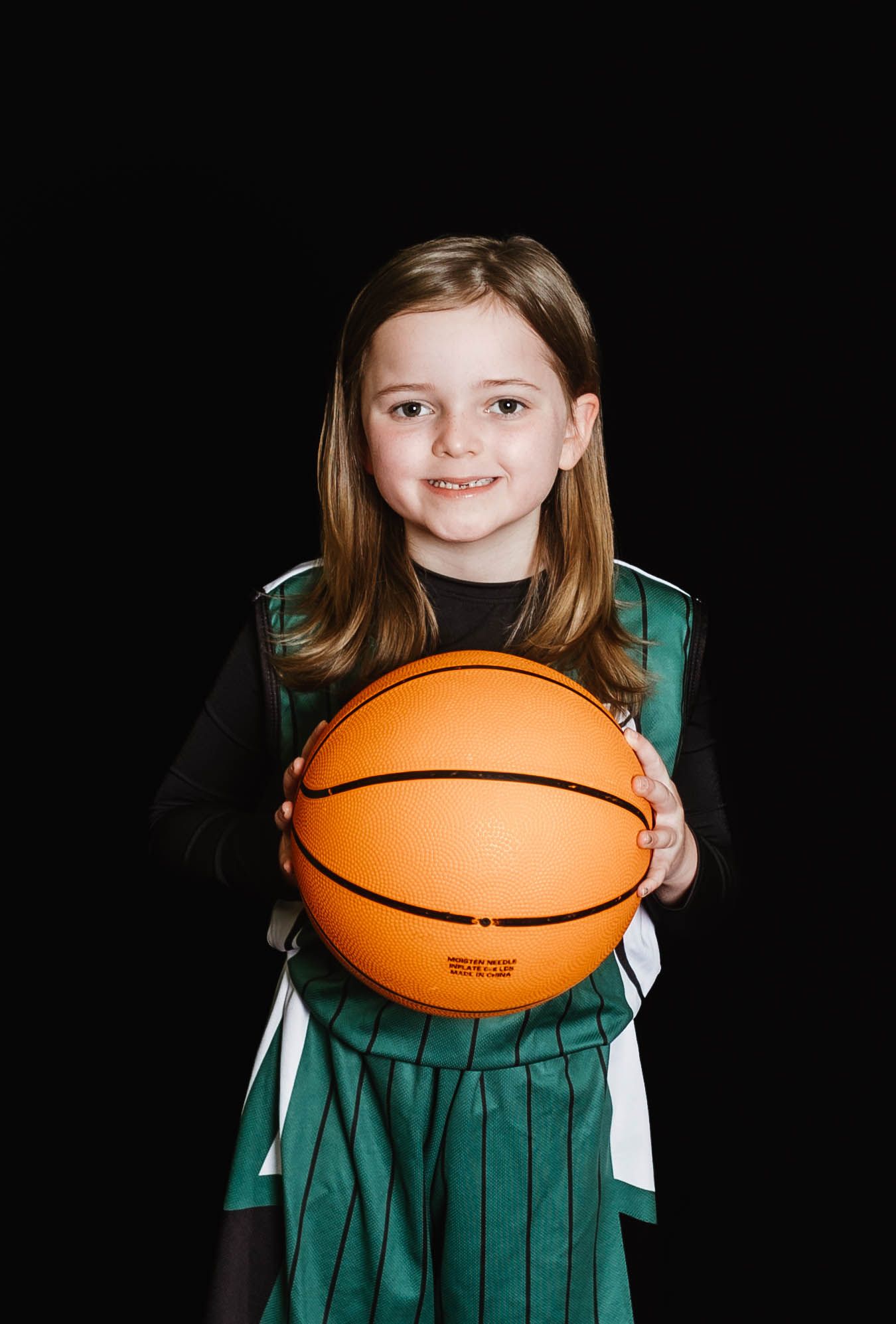 Youth basketball player in green and white uniform holds orange basketball against black background.
