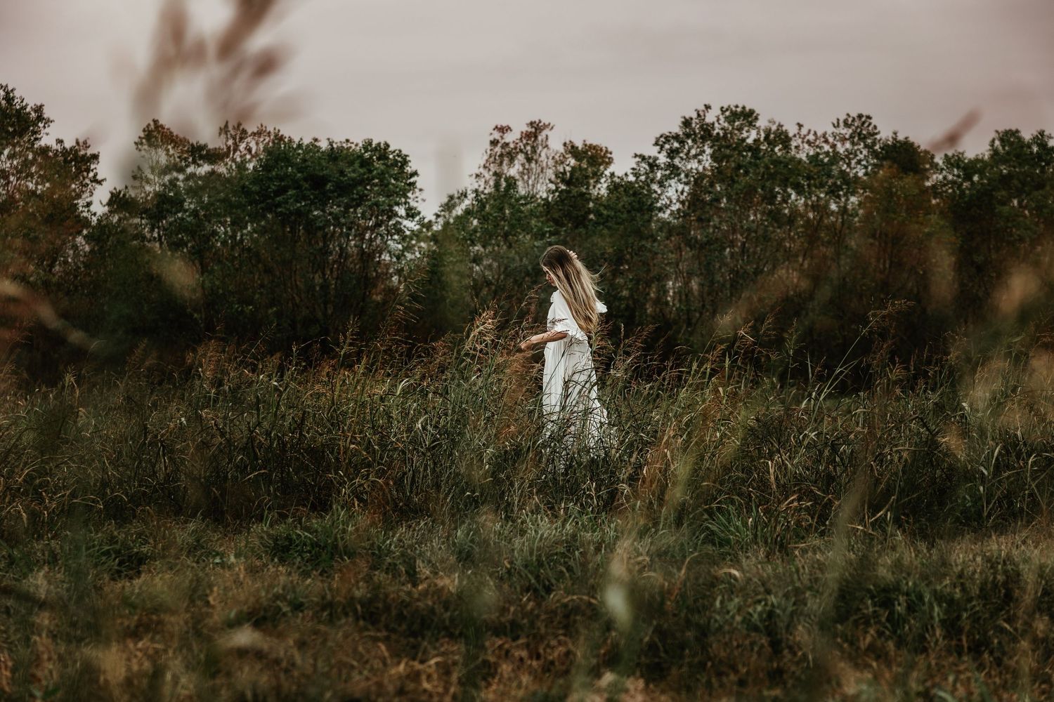 A figure in flowy white dress stands in a moody golden field surrounded by autumn trees at dusk.
