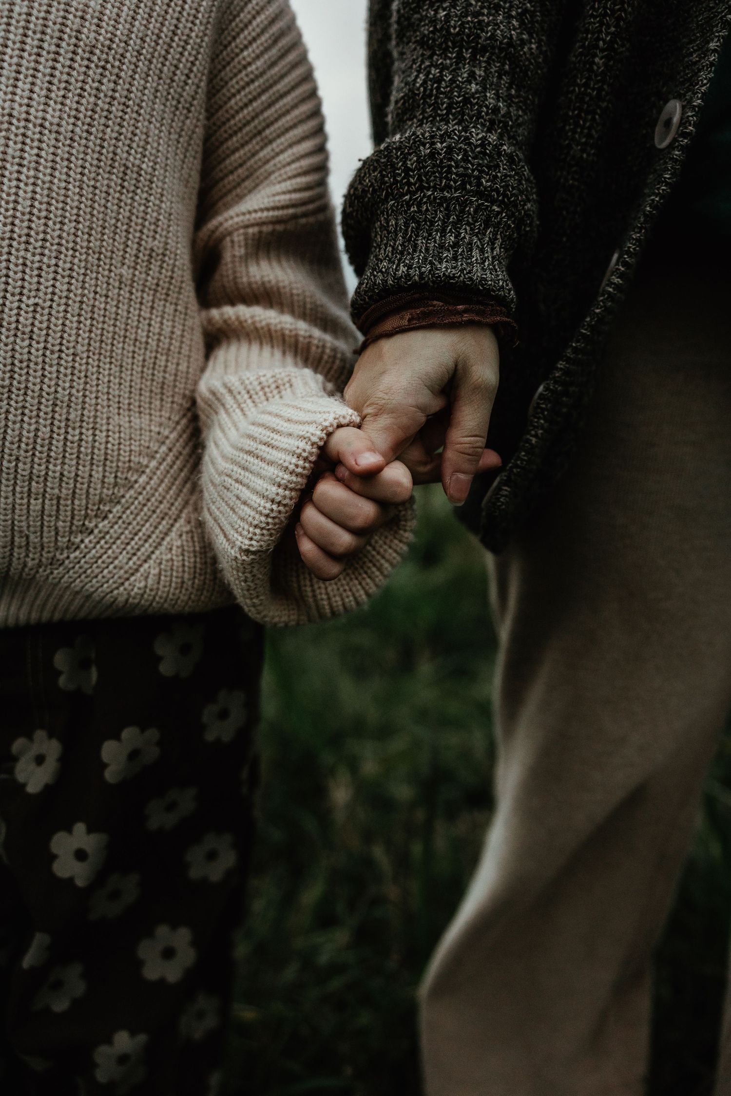 Close-up view of two people holding hands while wearing cozy knit sweaters against a floral and natural background.