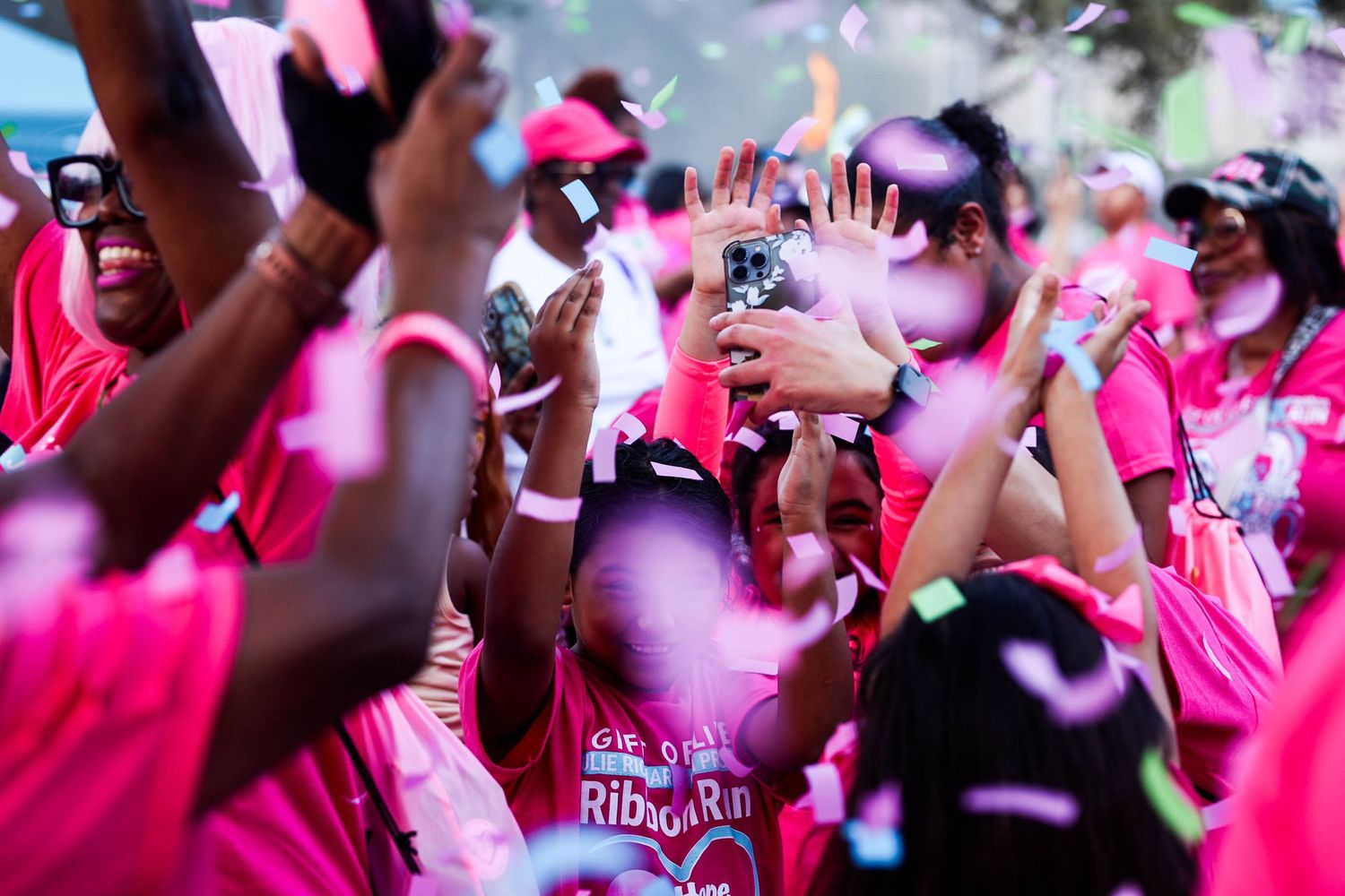 A crowd of participants dressed in pink celebrate at an outdoor event with confetti falling from above.