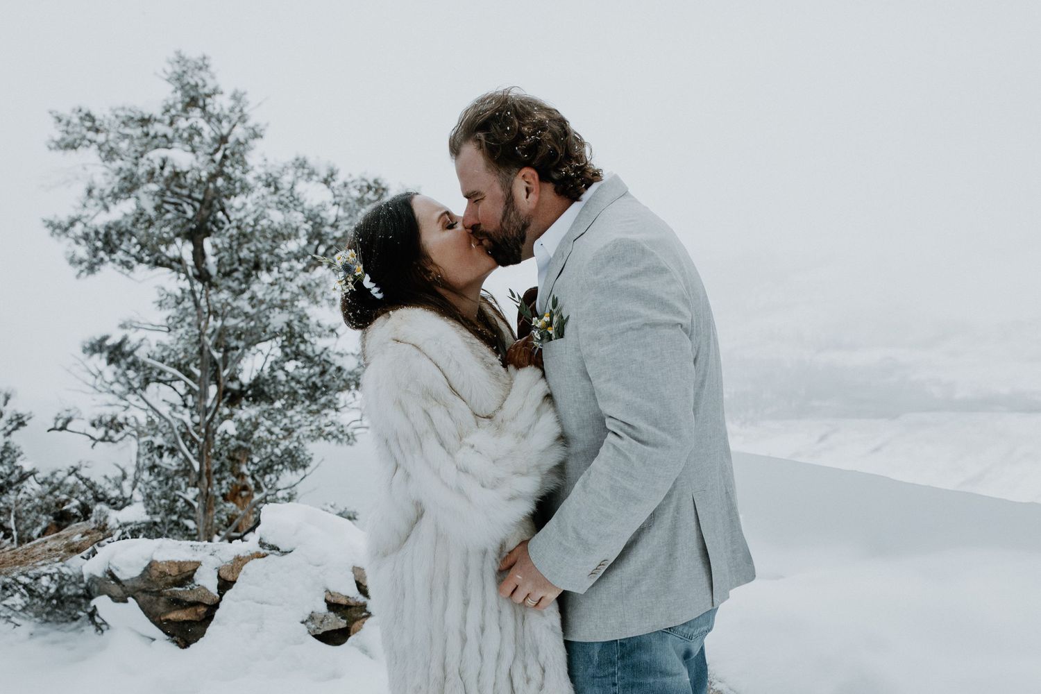A romantic winter wedding couple embraces in the snow with frost-covered evergreens in the background.