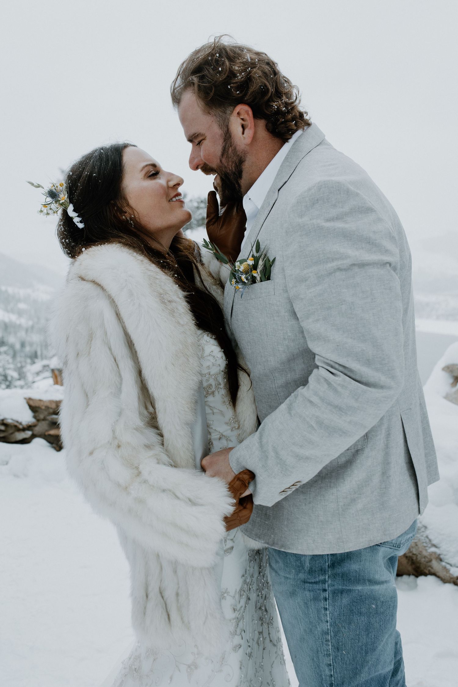 A couple shares an intimate winter moment in the snow, with the bride wearing a white fur coat and the groom in a gray blazer.