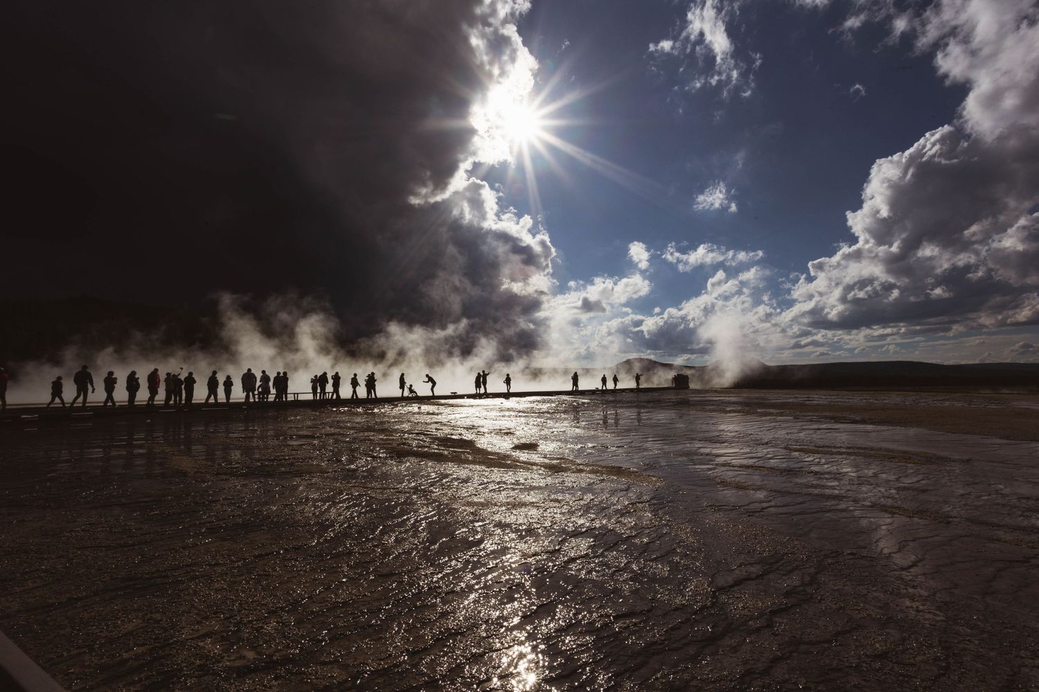 Silhouetted figures walk across a glistening landscape at Yellowstone's Grand Prismatic Spring during a sunny afternoon.