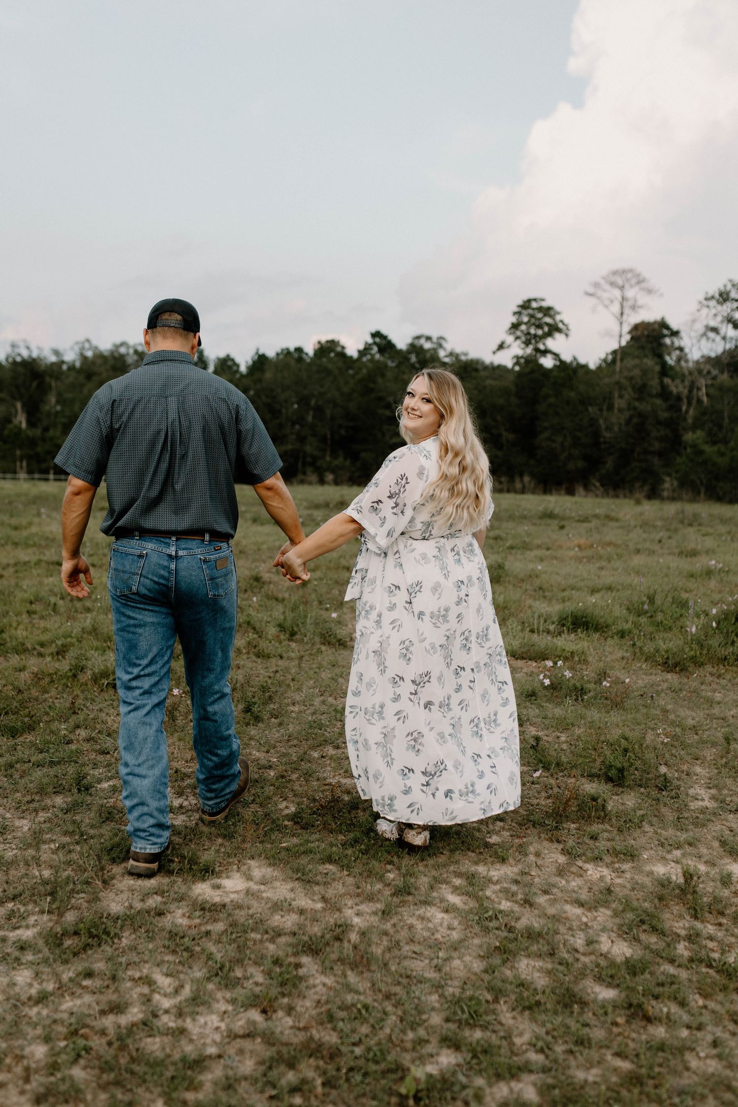 A couple in a field holding hands, with one wearing a floral dress and the other in jeans and a button-down shirt.