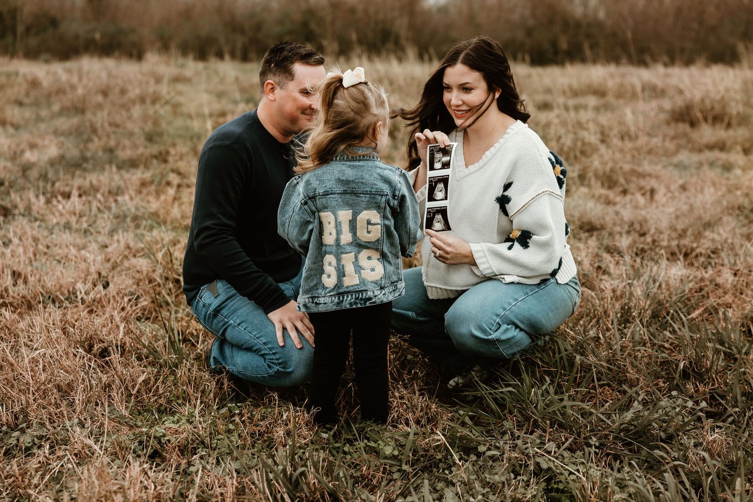 Family kneeling in autumn field wearing casual denim and sweaters during outdoor photo session.