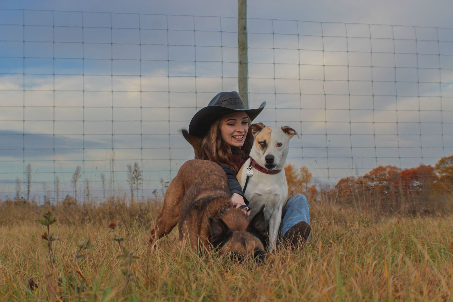 A woman in a cowboy hat smiles while sitting in a grassy field with two dogs near a fence.