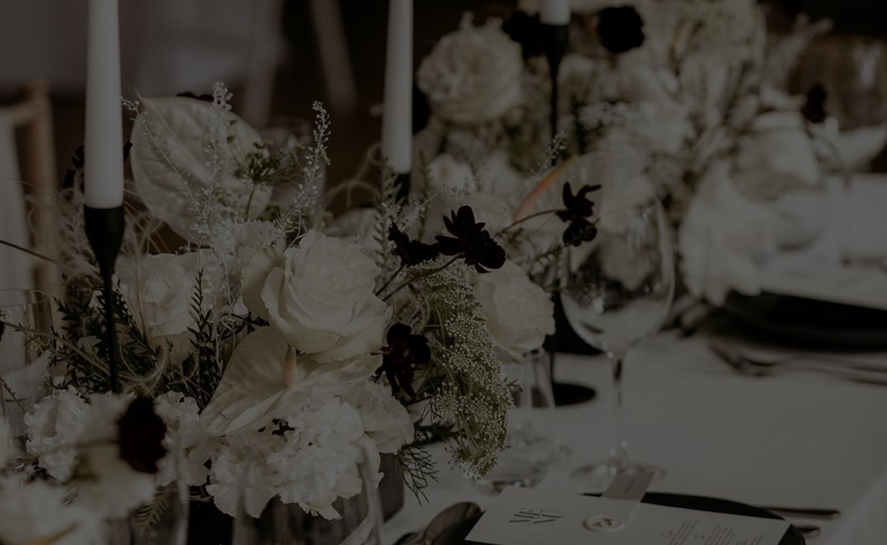 A black and white photograph shows elegant floral centerpieces with white roses on a formal dining table.
