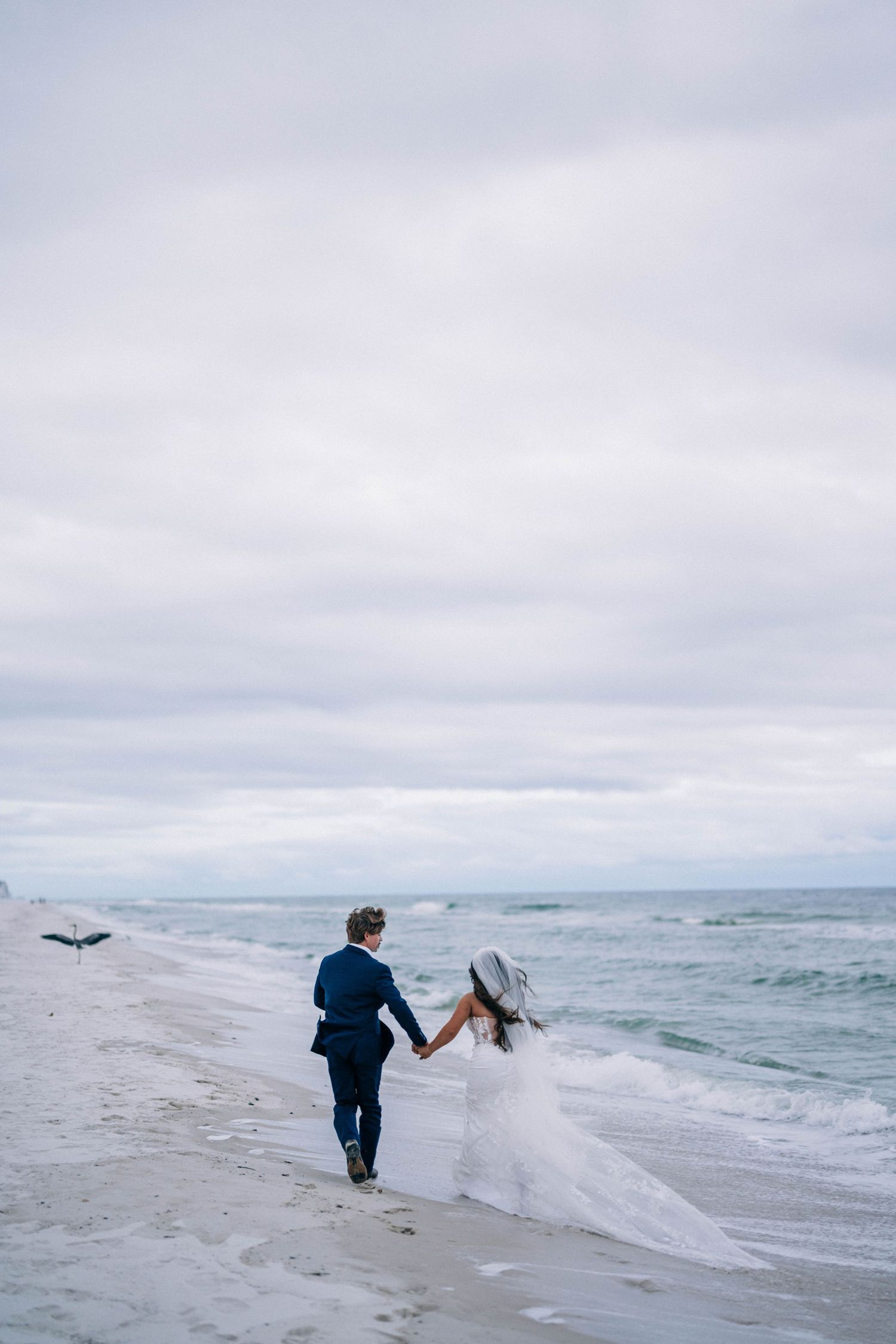 Bride and groom walk hand-in-hand along a sandy beach shoreline, with waves and overcast skies behind them.