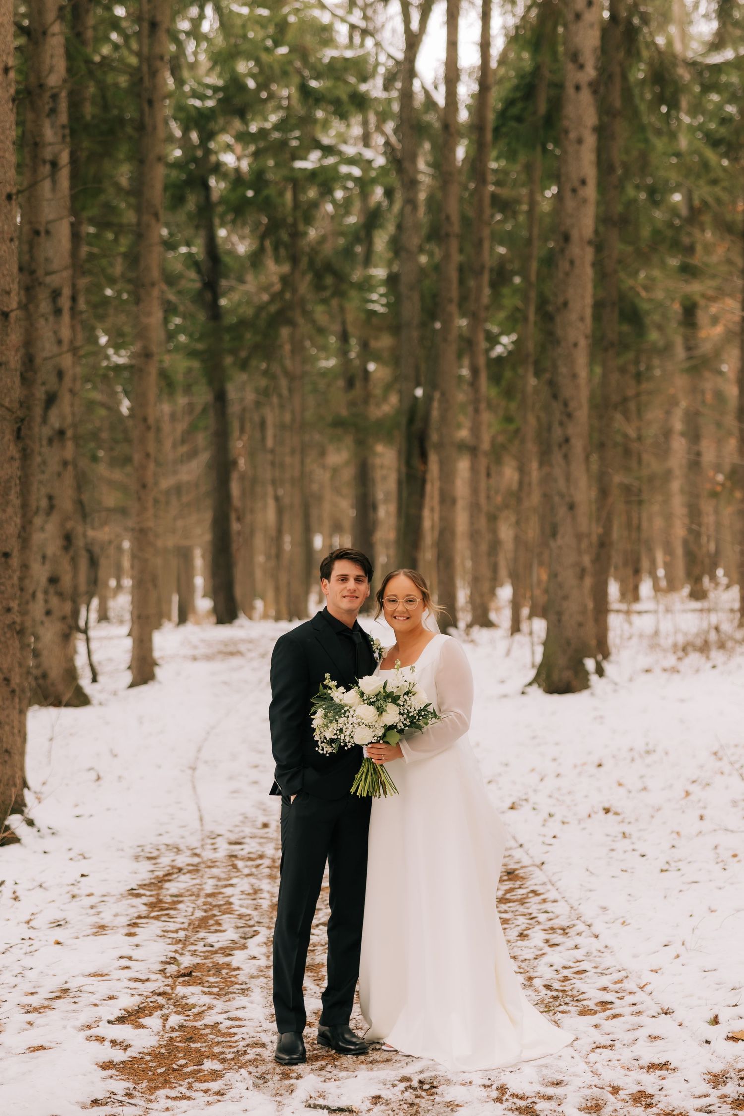 A couple poses together in a snowy winter forest wearing wedding attire with evergreen trees in the background.