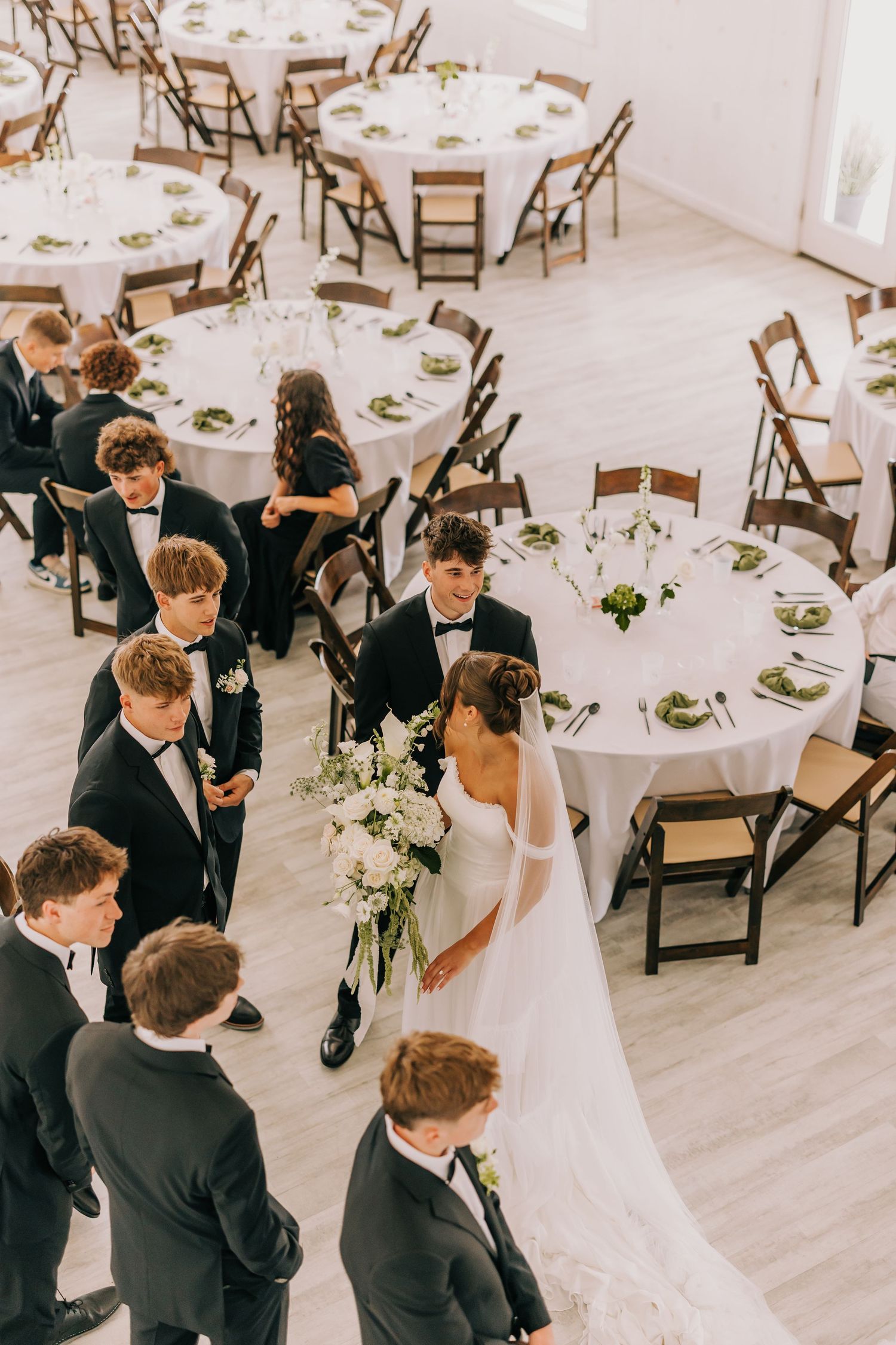 Elegant wedding reception scene with guests in formal attire gathering around tables in a bright white venue.