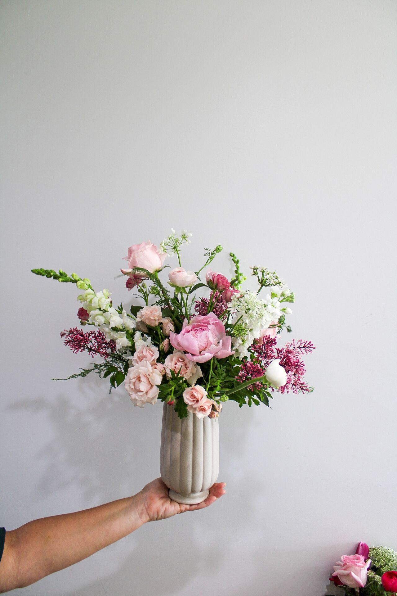 Hand holding a ceramic vase with a lush pink and white floral arrangement featuring peonies and roses against a gray wall.