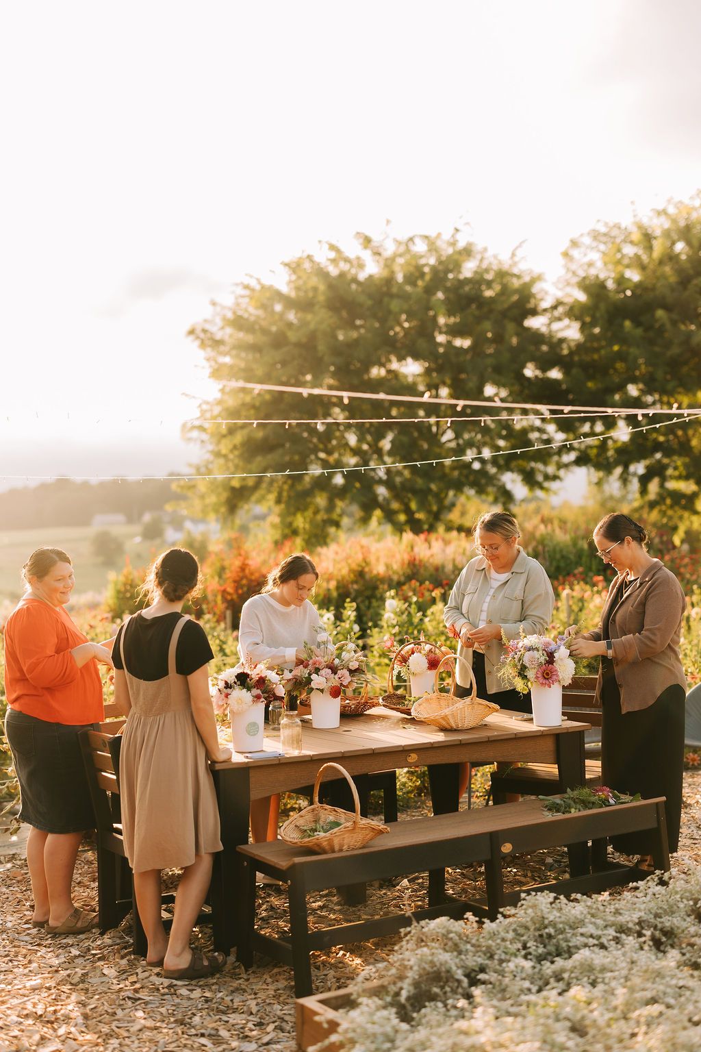 People gather at rustic outdoor dining tables during golden hour sunset with white floral arrangements and natural decor.