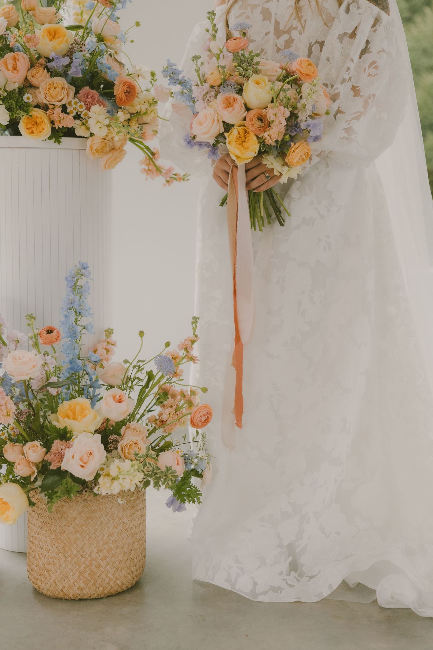 Pastel wedding bouquets with peach roses and blue flowers arranged with flowing ribbons against white fabric.
