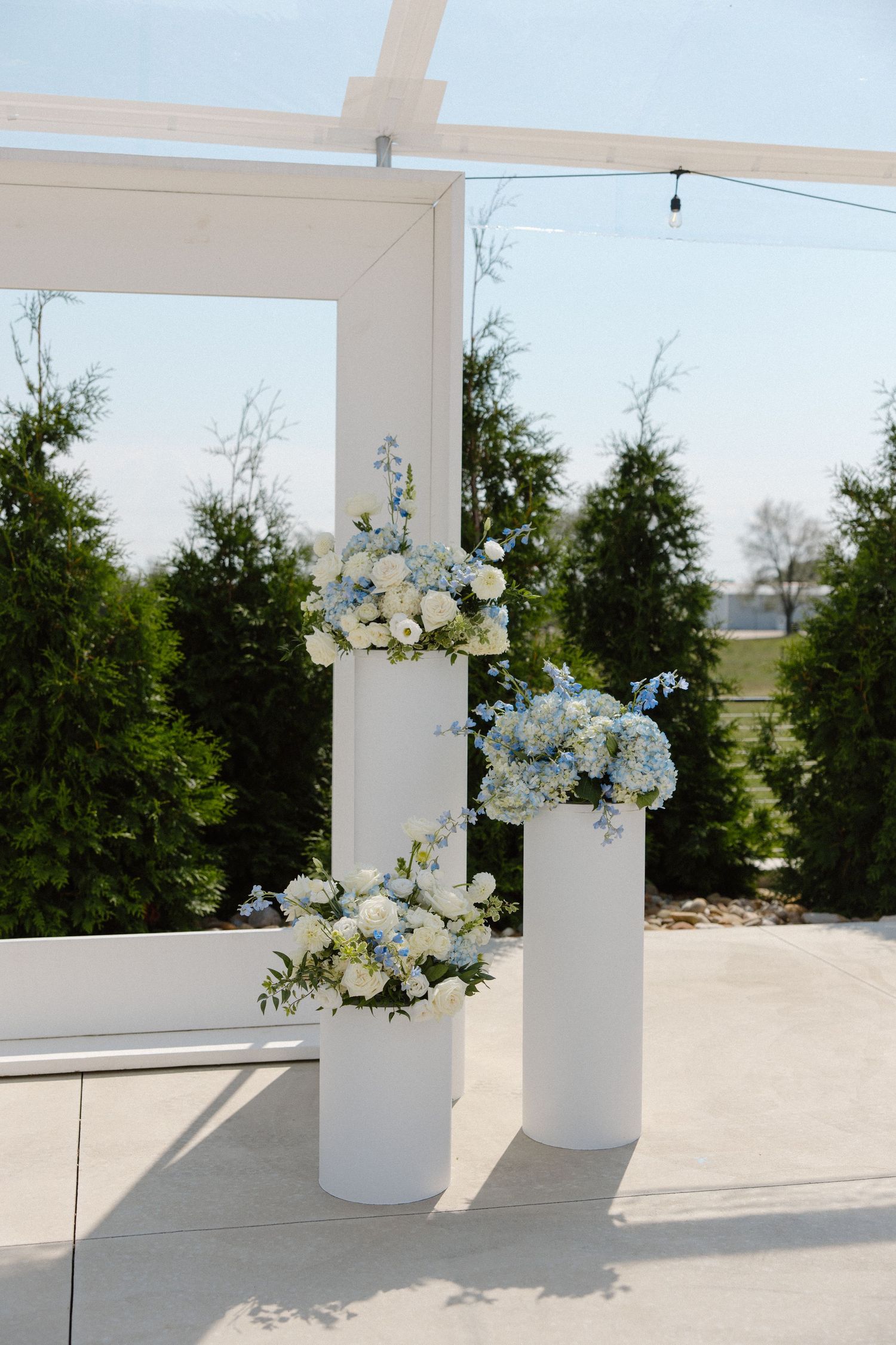 Modern white pedestals with blue and white floral arrangements against evergreen backdrop.
